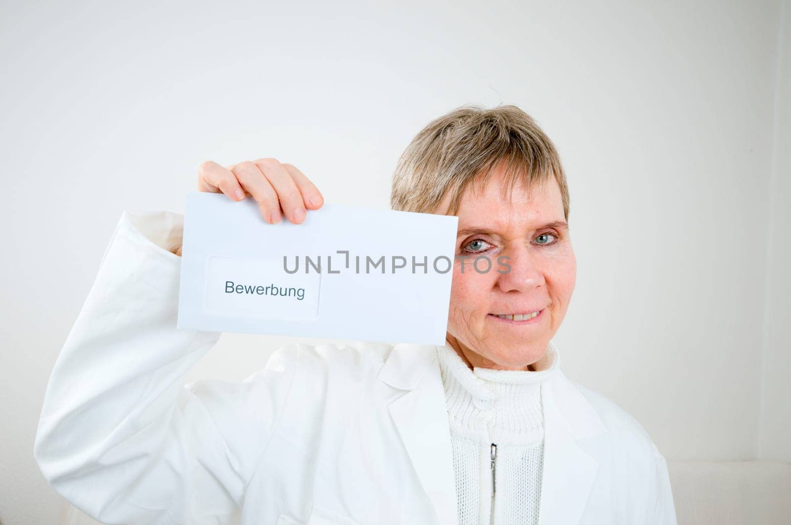 Upper body shot of an elderly woman in a white doctor's coat holds up an envelope with application inscription next to her