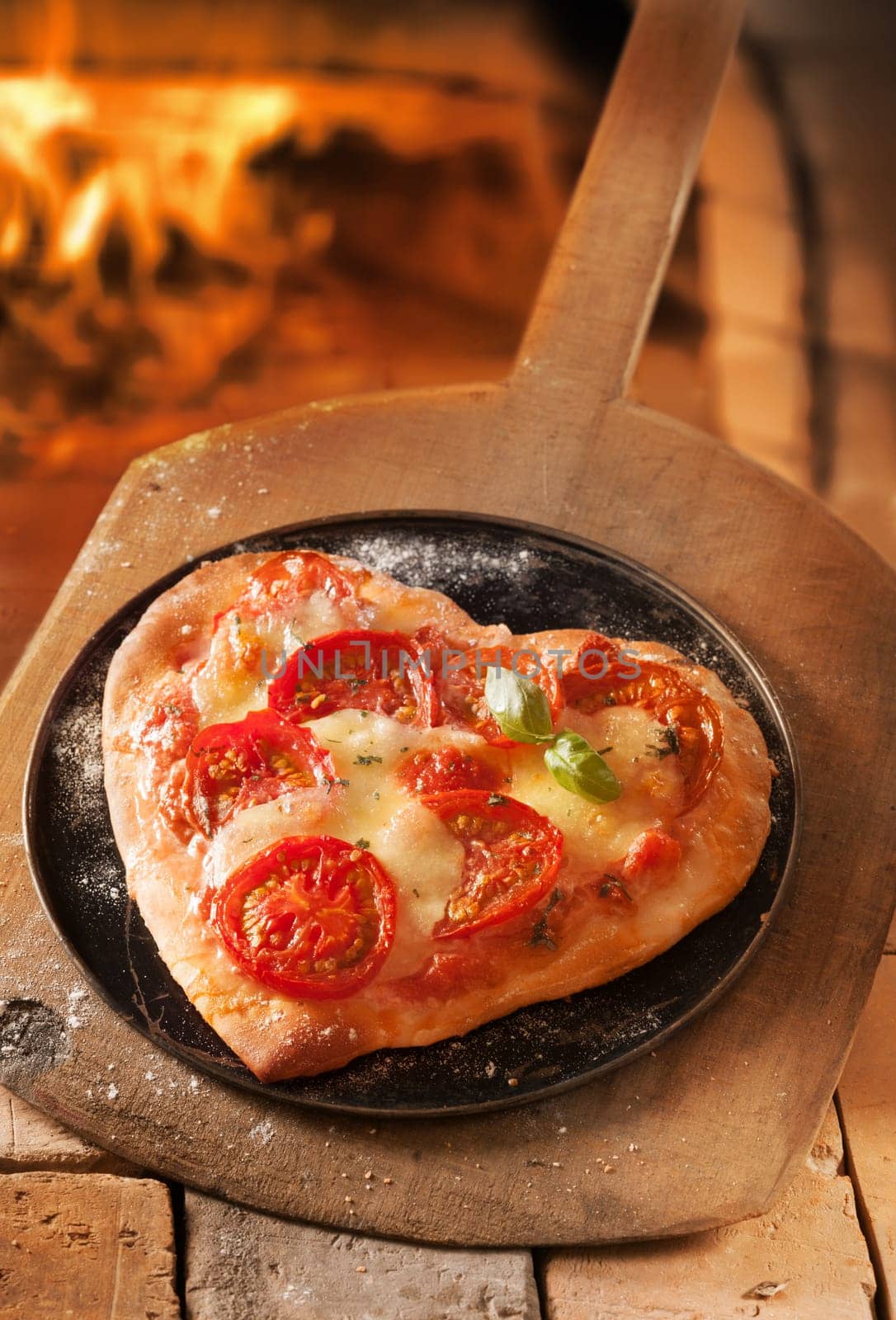 Romantic heart shaped Italian pizza on a platter and wooden board with the fire of a pizza oven visible behind