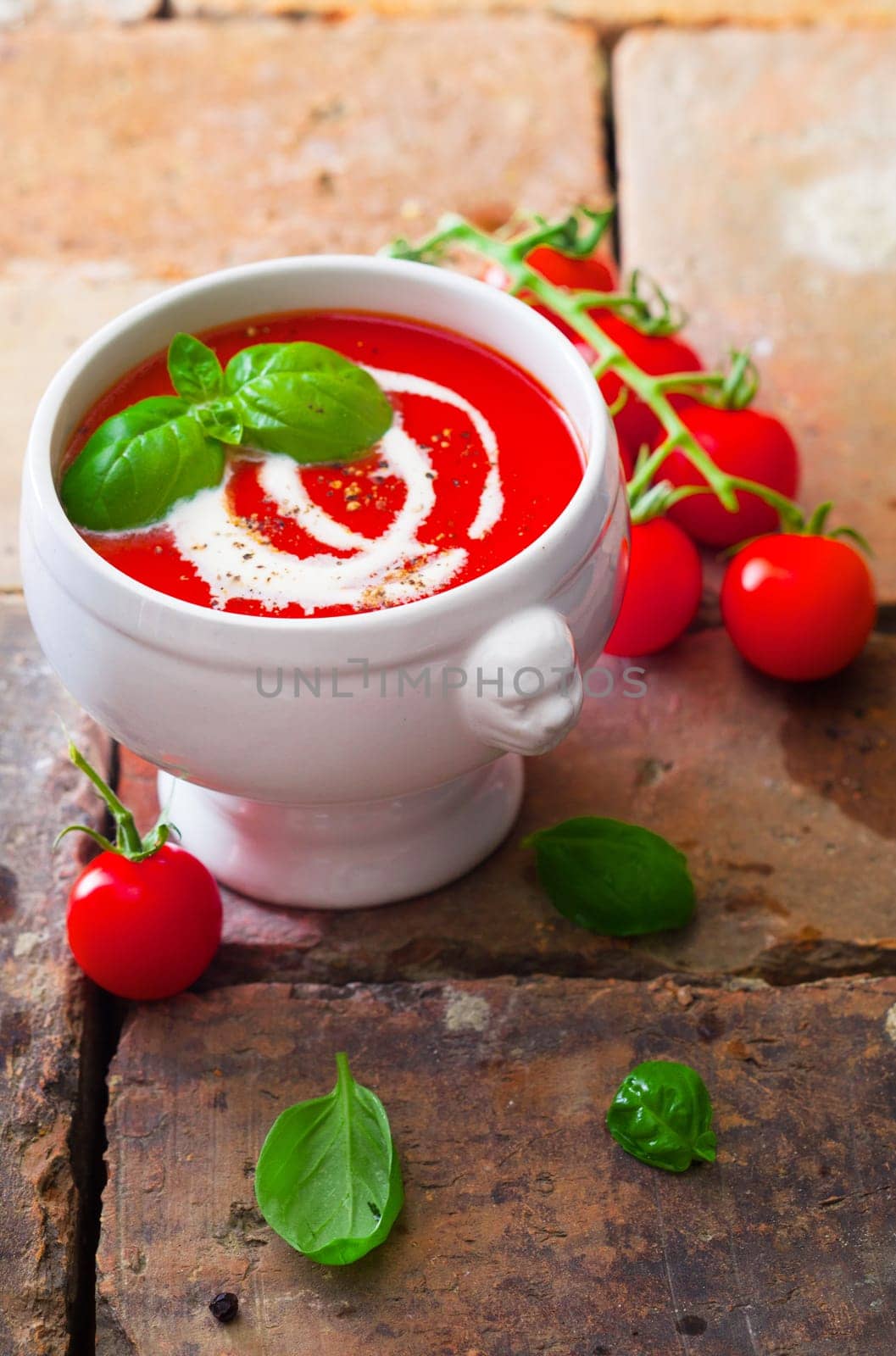 Small bowl of colourful red tomato soup with cream and basil on old weathered bricks with scattered fresh ingredients