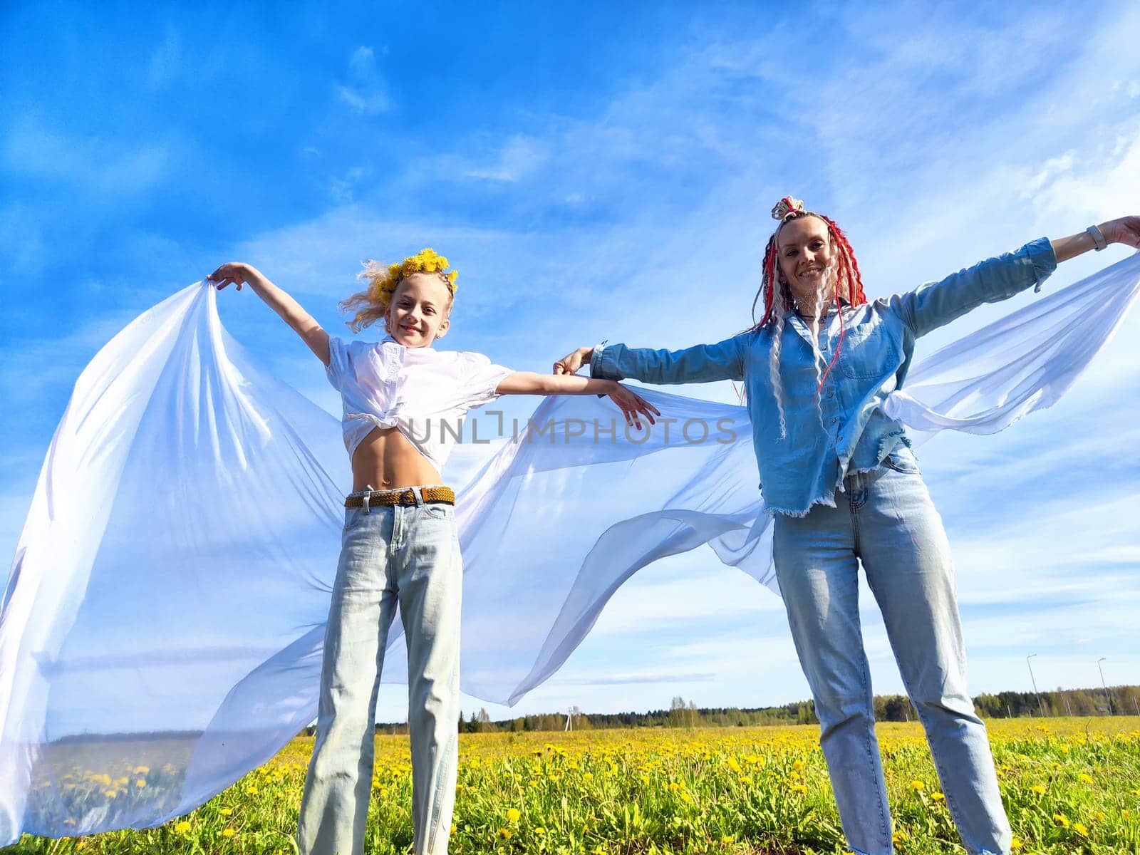 Two children joyfully wave white fabric while standing in a sunny field filled with bright yellow flowers under a clear blue sky.