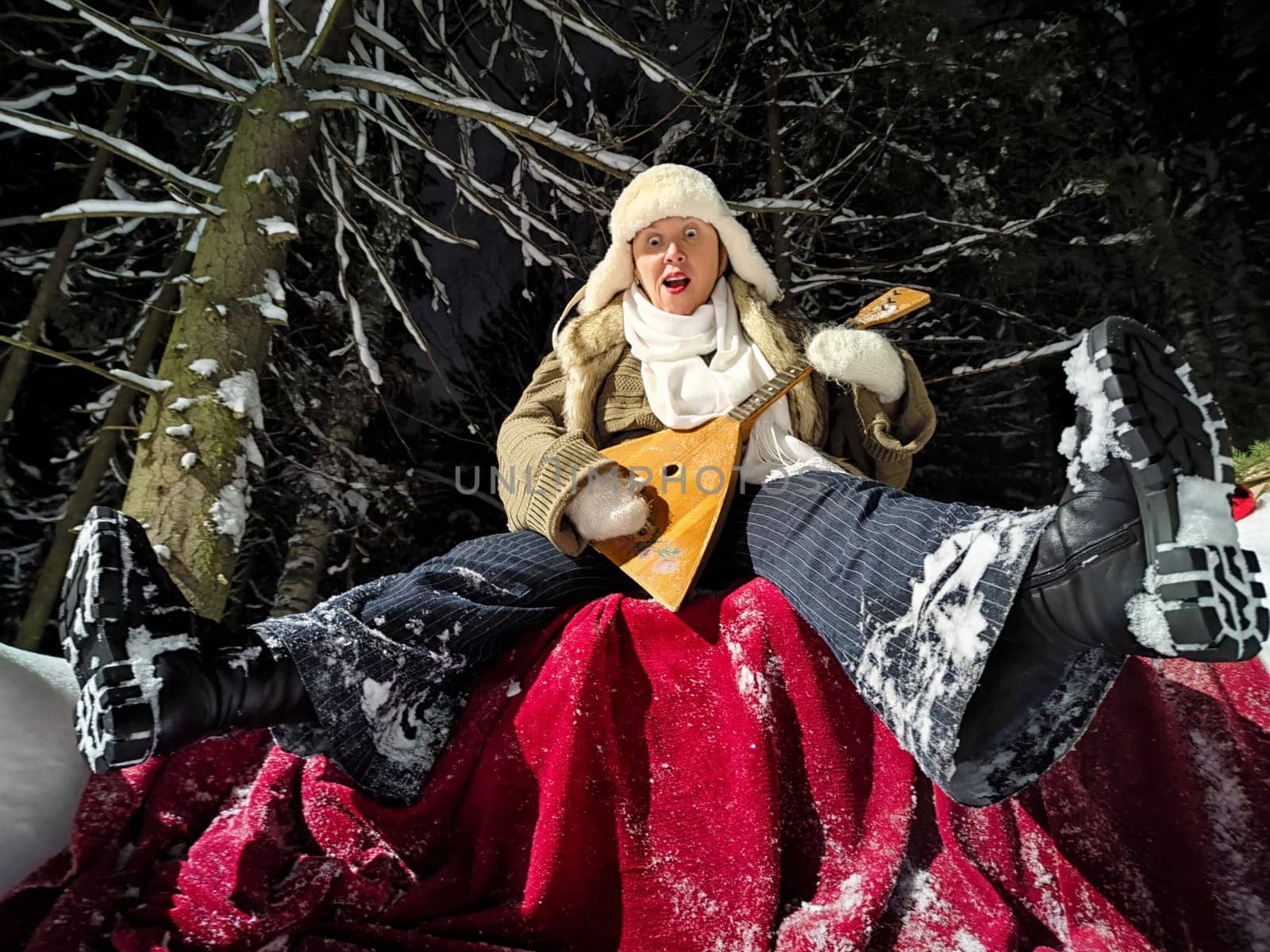 A middle-aged woman with a funny expression walks through a snowy forest at night, wearing a warm hat and playing a balalaika.