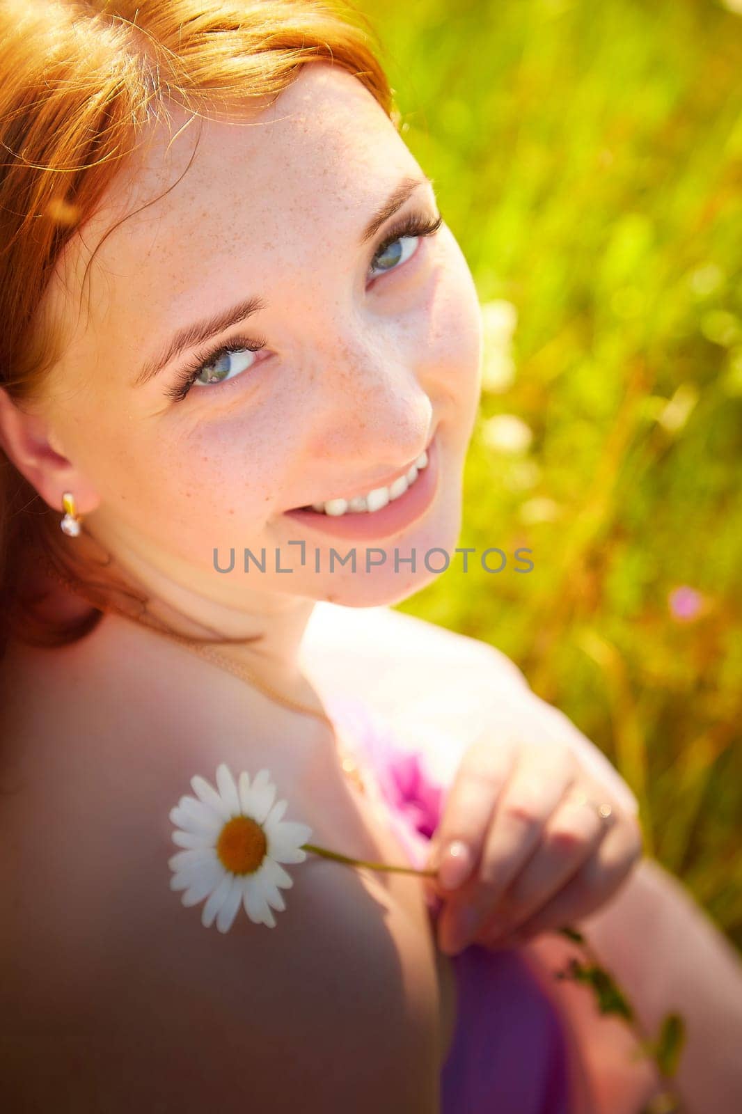 Cheerful young woman with red hair holds a daisy, surrounded by vibrant green grass and wildflowers on a sunny summer day.