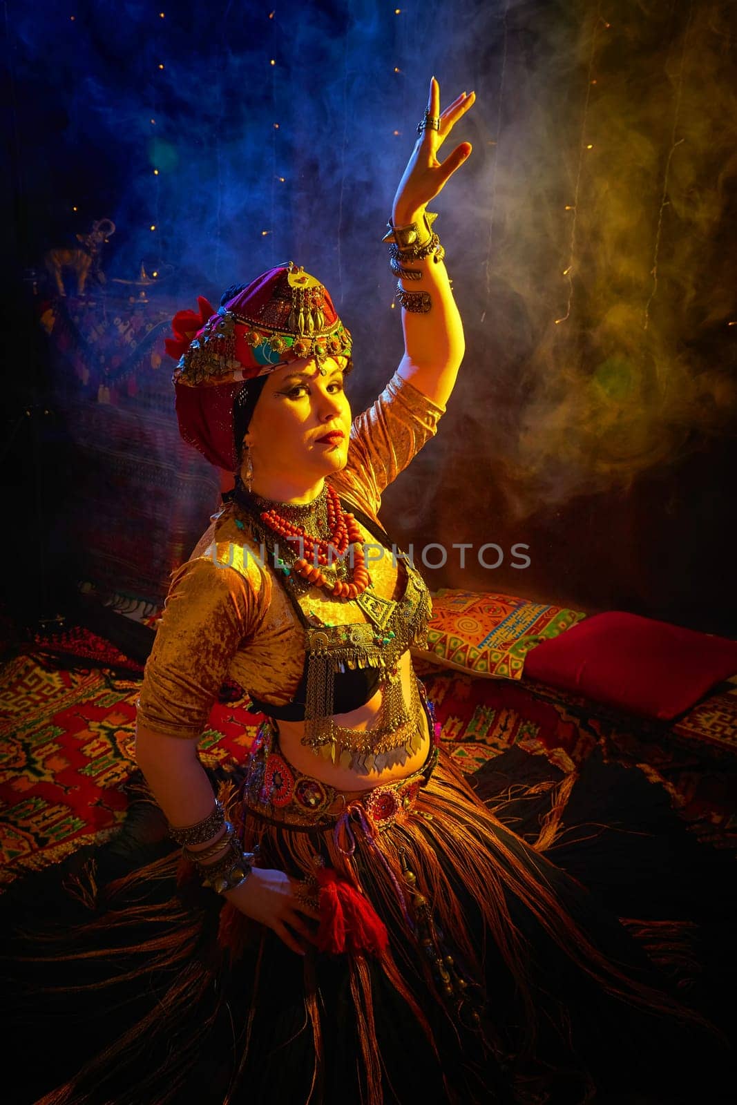 A woman in a colorful headscarf and jewelry poses in front of wall of colorful tassels