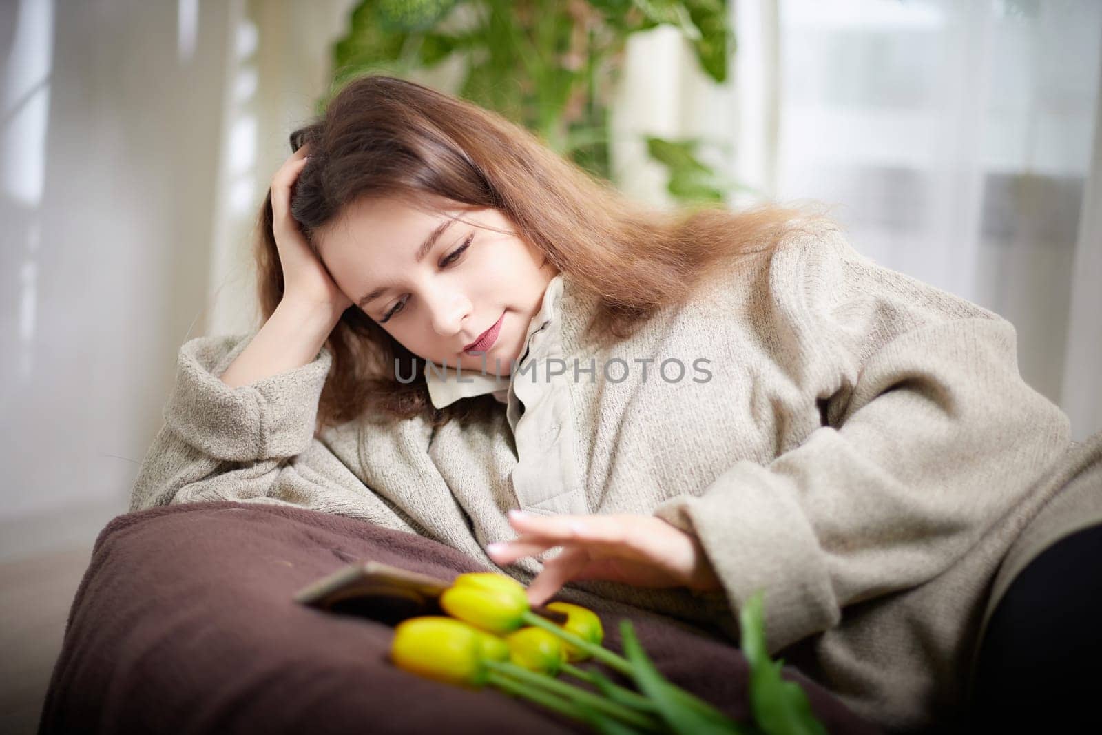 A woman comfortably lies on a couch, engaged with her smartphone while beautiful flowers add a touch of nature to her relaxing space.