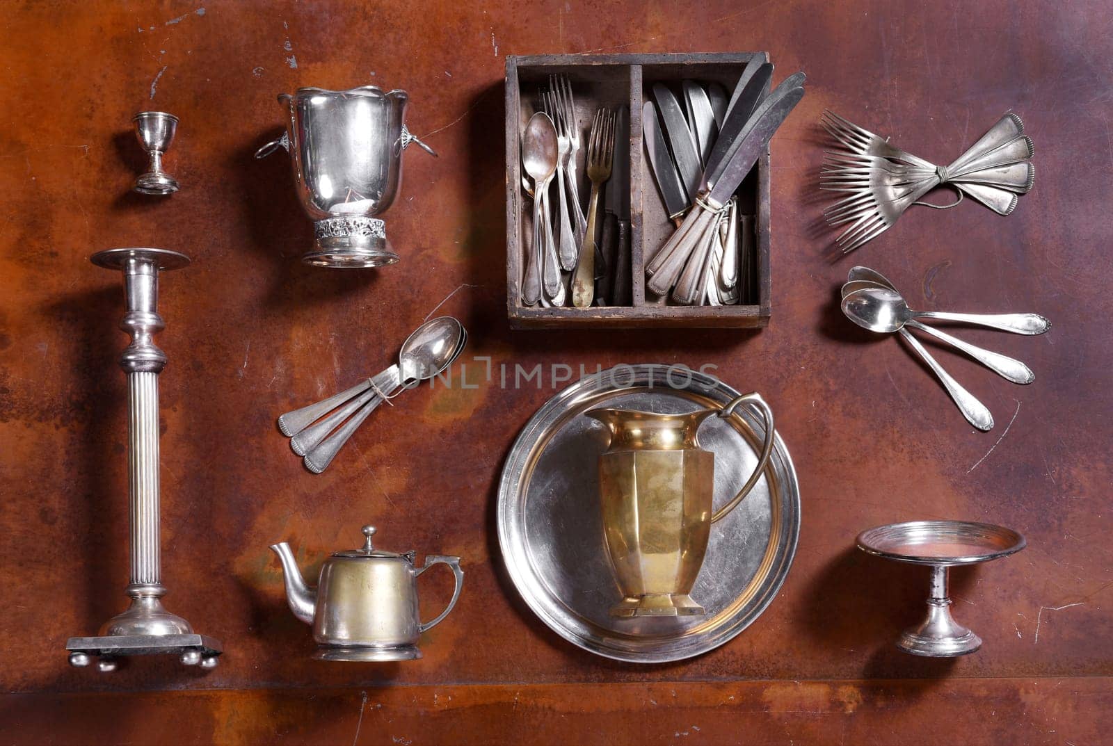 Assorted silver kitchenware arranged on wood with cutlery, candlestick, ice bucket, egg cup, teapot tray and pitcher or jug viewed from above