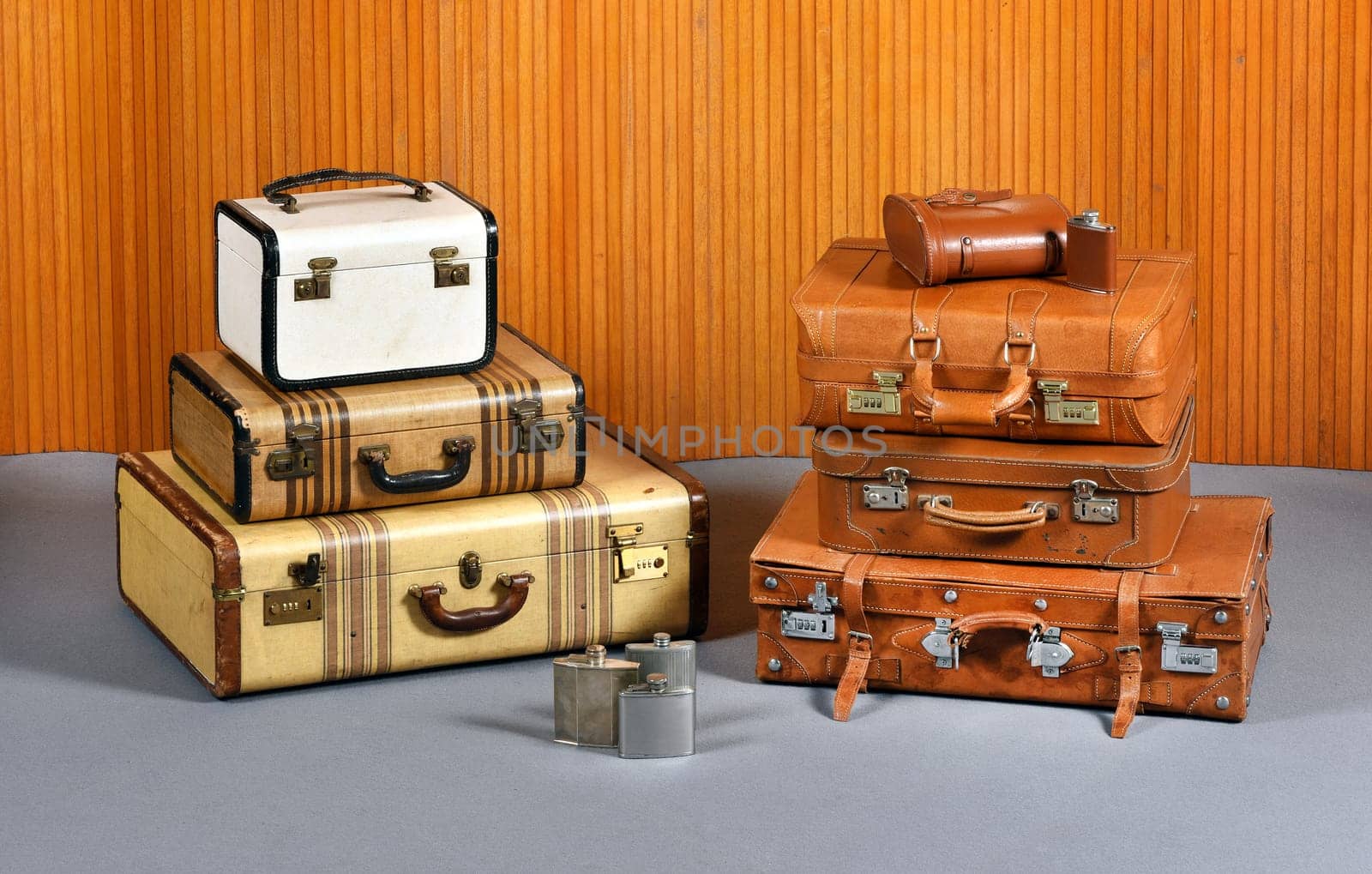 Still Life of Antique Suitcases and Flasks - Two Stacks of Various Sized Suitcases and Trunks with Collection of Metal Alcohol Drinking Flasks in Room with Gray Floor and Wood Wall Paneling