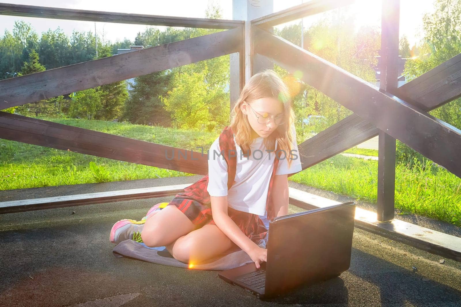 A young girl sits on the ground with a laptop, focused on her work in a sunny outdoor location surrounded by greenery.