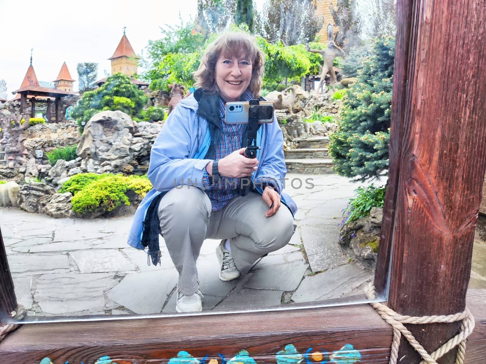 Middle-aged woman takes a humorous selfie in a warped mirror surrounded by lush greenery in a park setting.