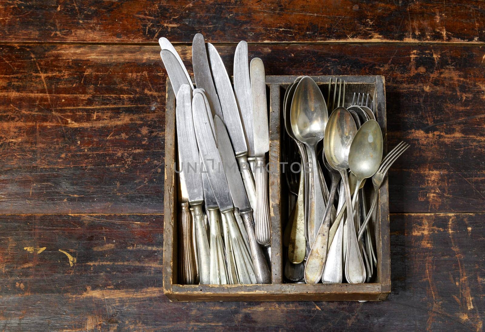 Collection of old vintage cutlery in a wooden tray with assorted silver spoons, knives and forks in an overhead view on an old rustic wooden board