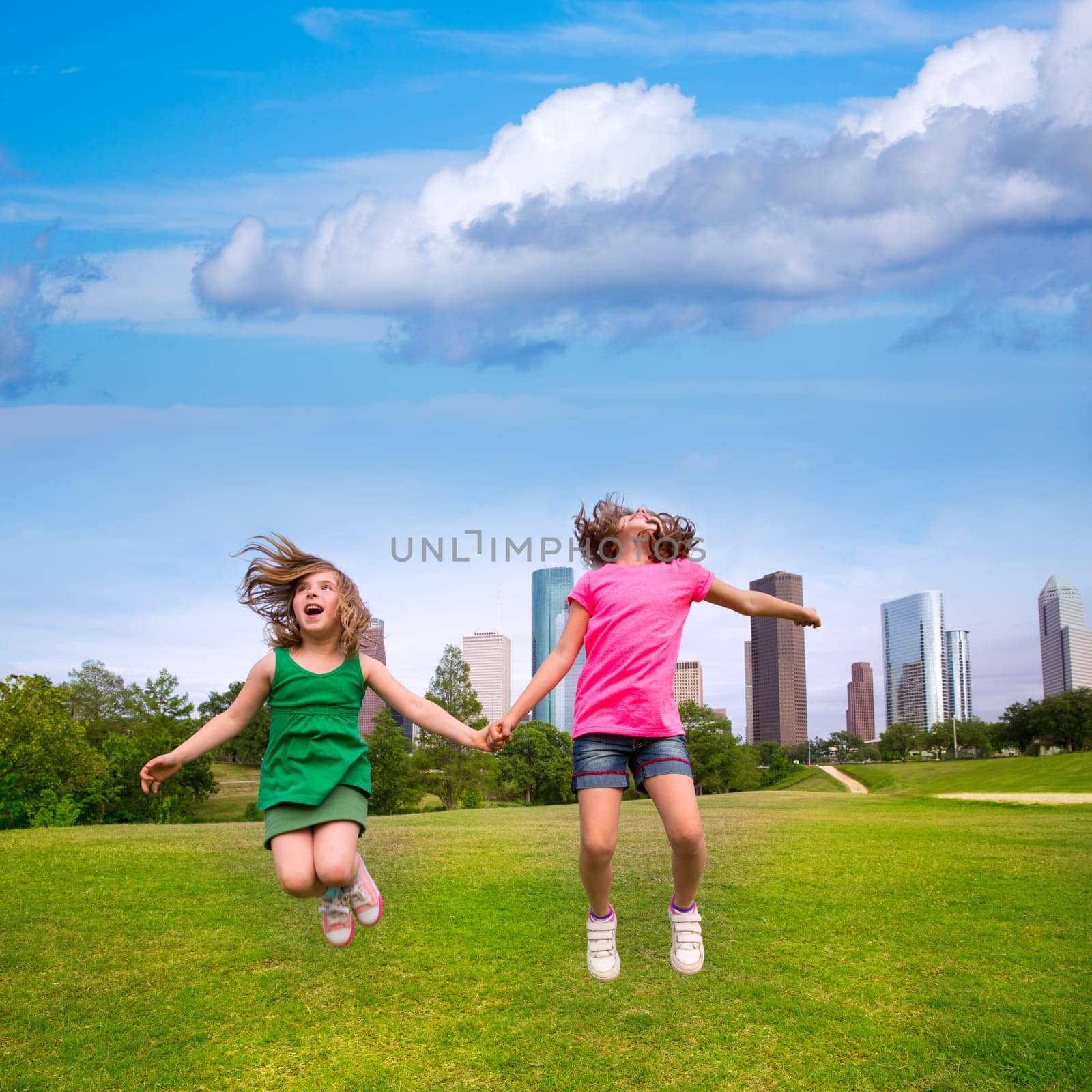Two sister girls friends jumping happy holding hand in urban modern skyline on park lawn