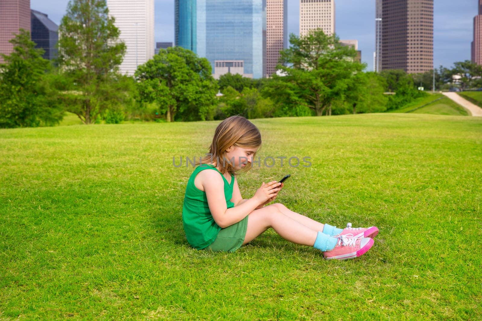 Blond kid girl playing with smartphone sitting on park lawn in city skyline background