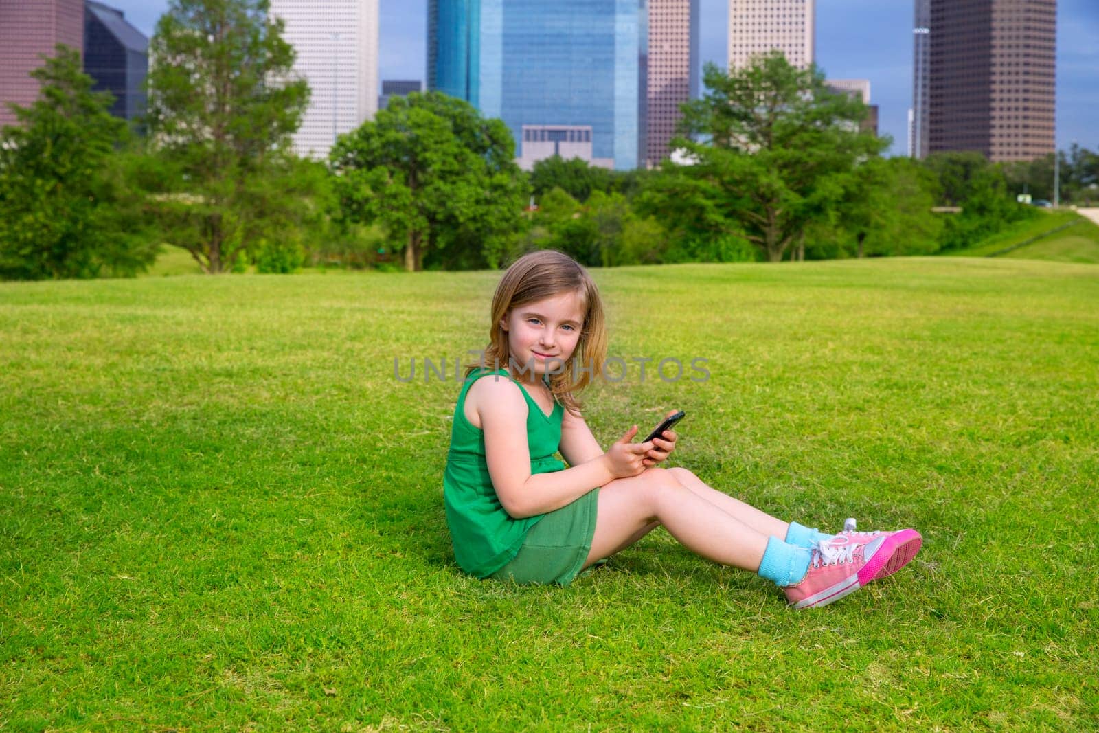 Blond kid girl playing with smartphone sitting on park lawn in city skyline background
