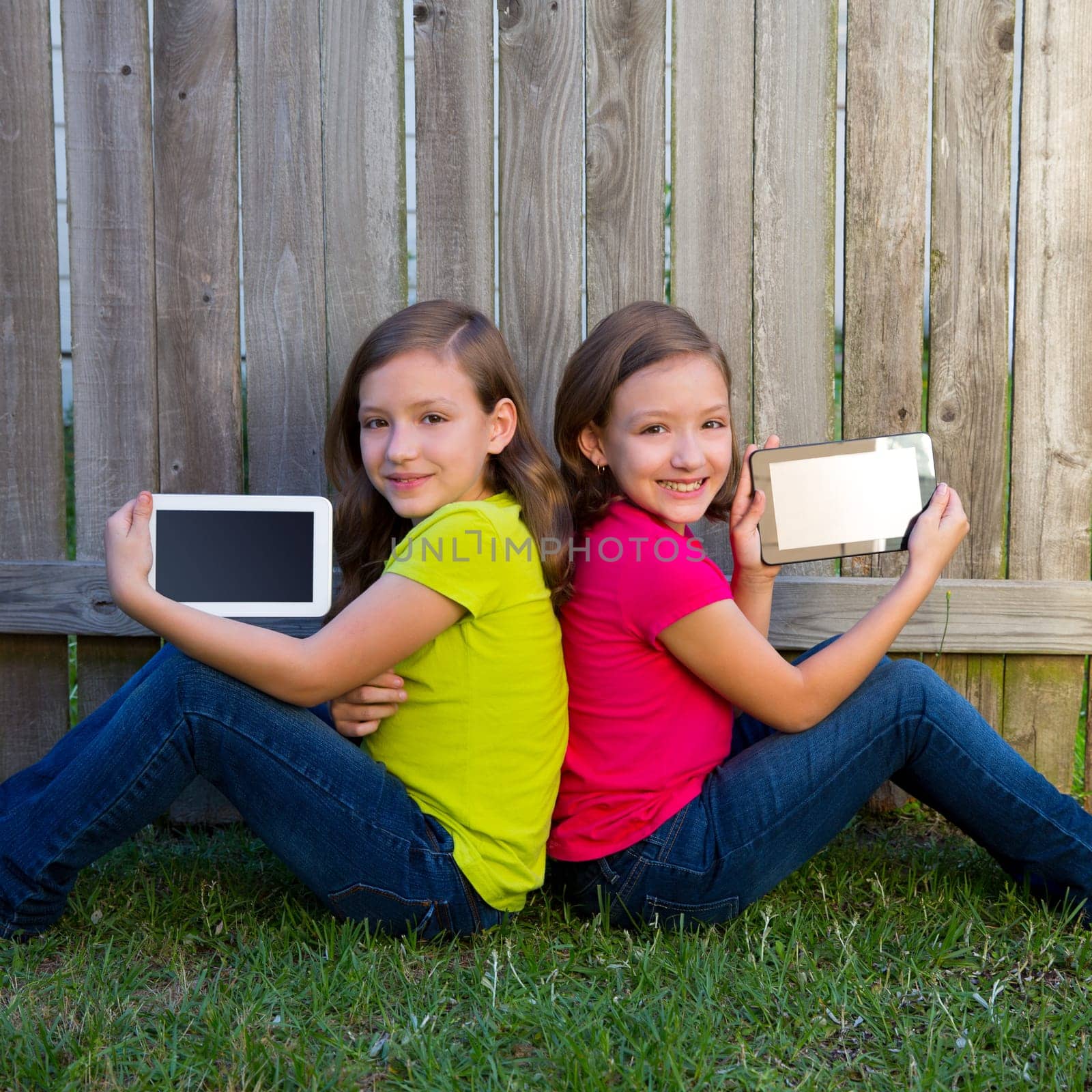 Twin sister girls playing with tablet pc sitting on backyard lawn fence leaning on her back