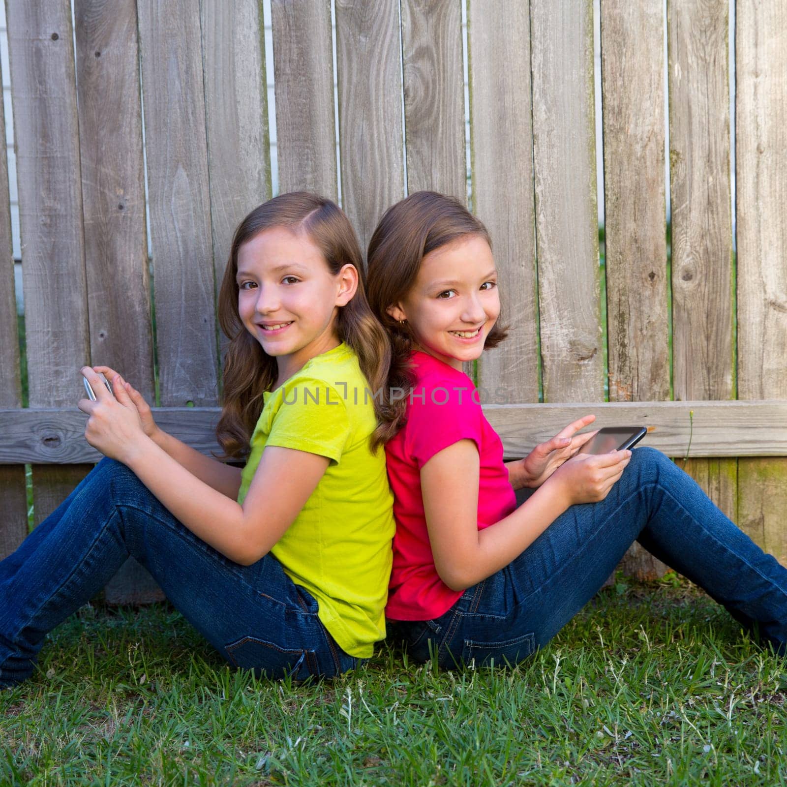 Twin sister girls playing with tablet pc sitting on backyard lawn fence leaning on her back