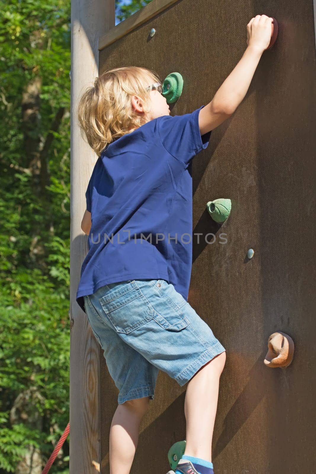 The blond boy climbs the climbing wall in summer day.