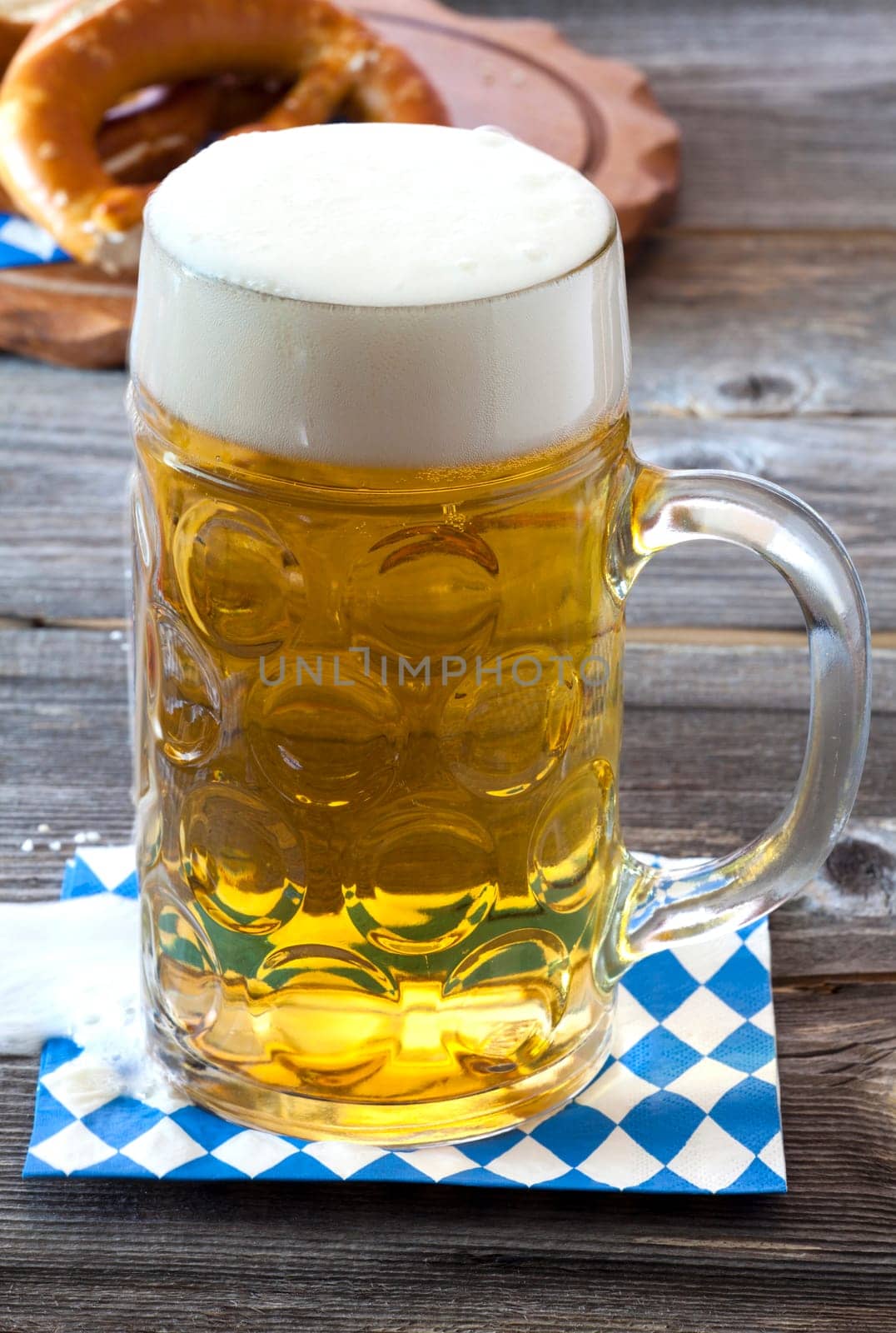 Large cool glass of beer on a napkin with blue and white diamond pattern on an old rustic wooden table in the background a wooden board with pretzels