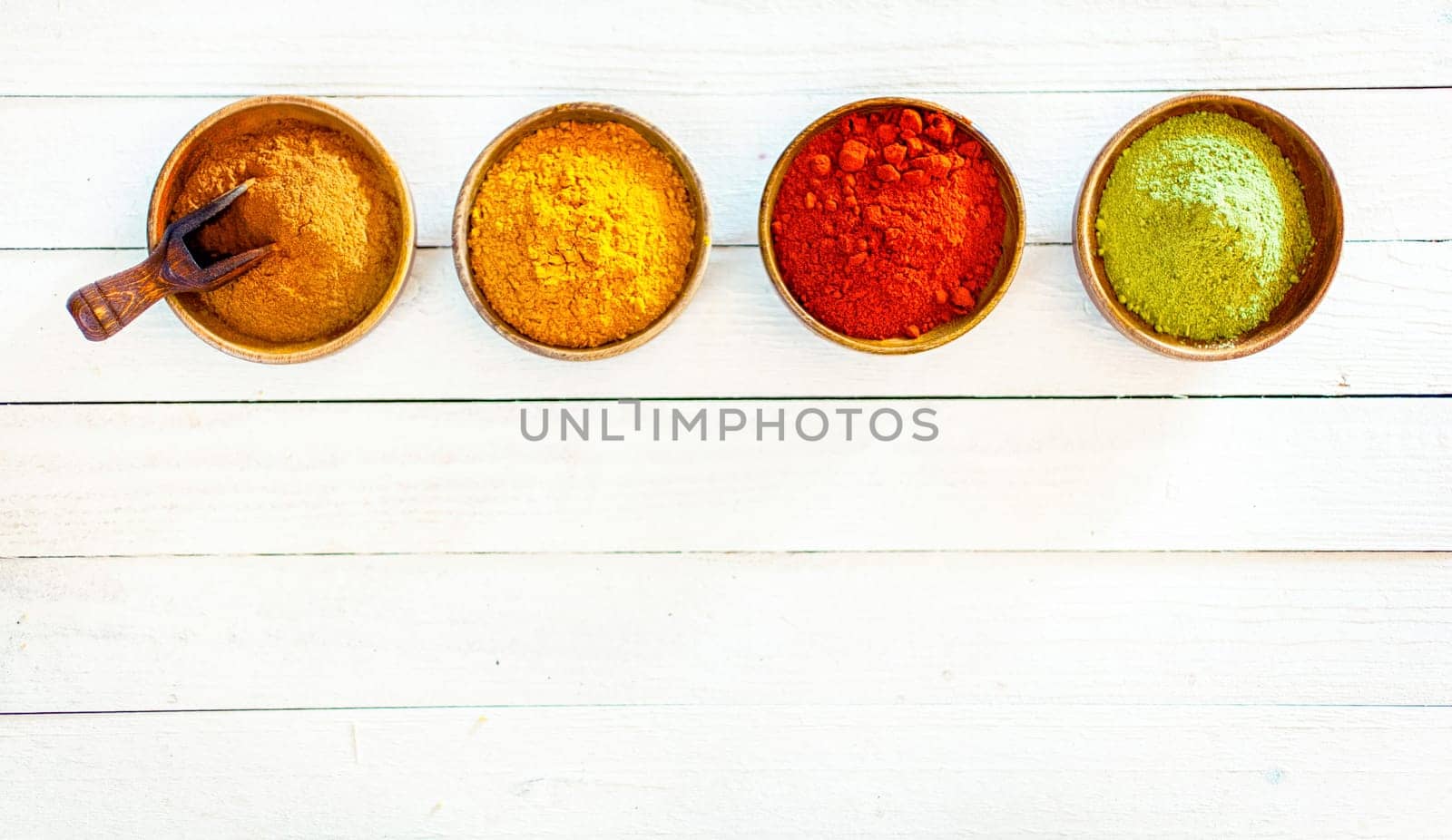 Overhead view of a line of four colourful ground spices in wooden bowls on white painted boards with copyspace below