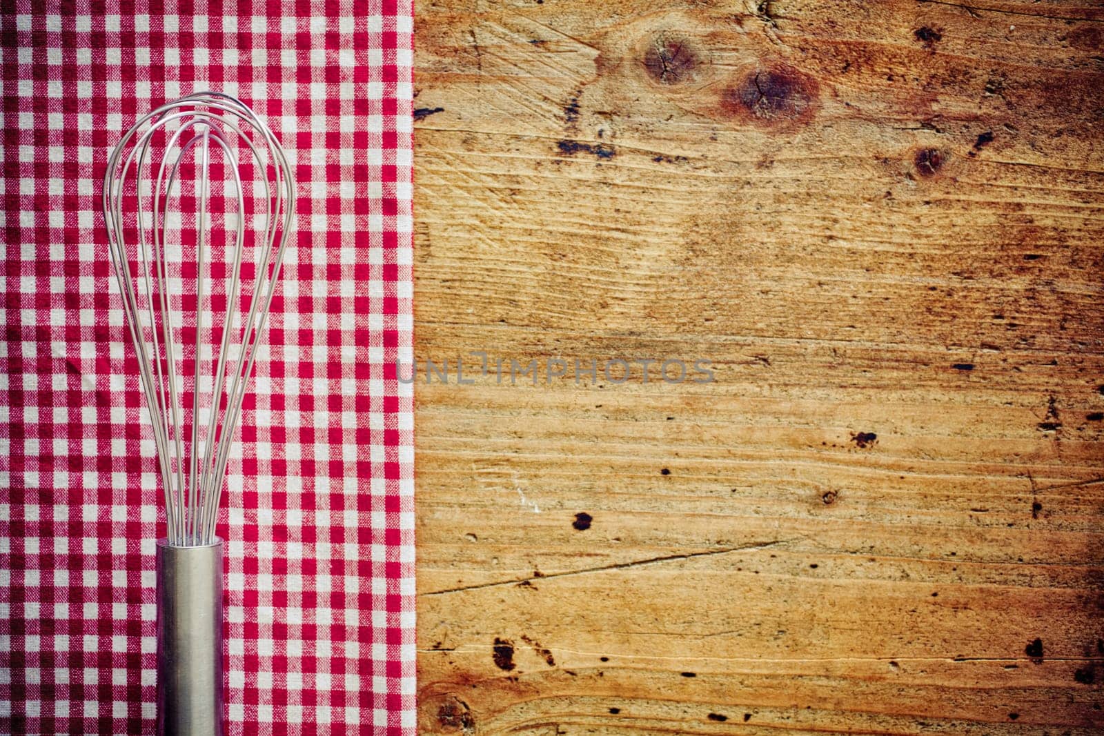 Metal cooking whisk, or balloon whisk, lying on a fresh red and white checkered napkin on a wooden countertop in a rustic kitchen with copyspace
