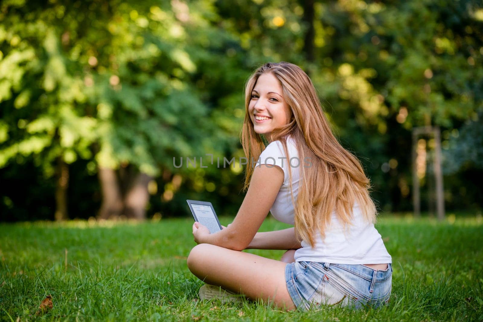 Young woman (teen girl) reading book on electronic book reader - outdoor, rear view