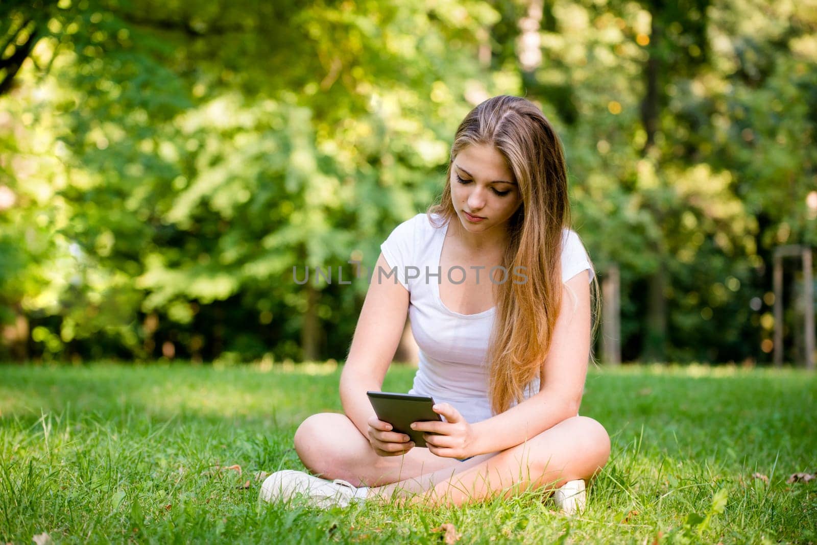 Young woman (teen girl) reading book on electronic book reader - outdoor in nature