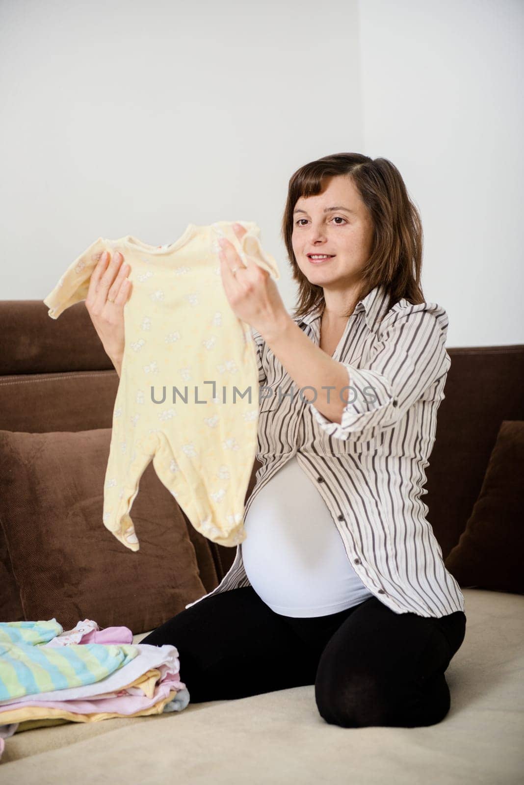 Pregnant woman preparing baby clothes at home on sofa