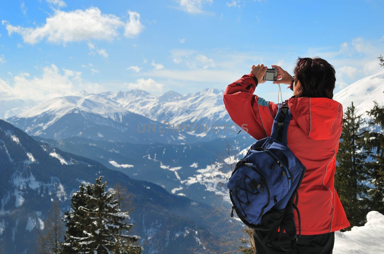 Senior woman with backpack holding camera and taking picture in winter mountains - rear view
