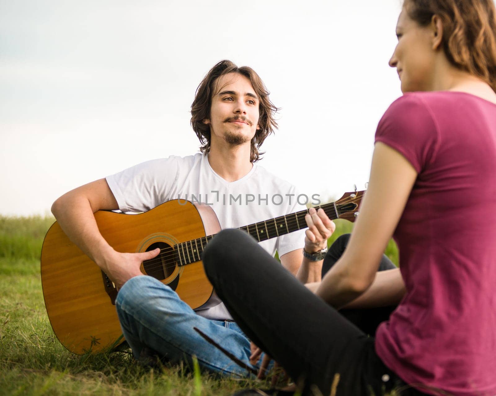 Young man playing to his girlfriend on guitar in nature - outdoor lifestyle