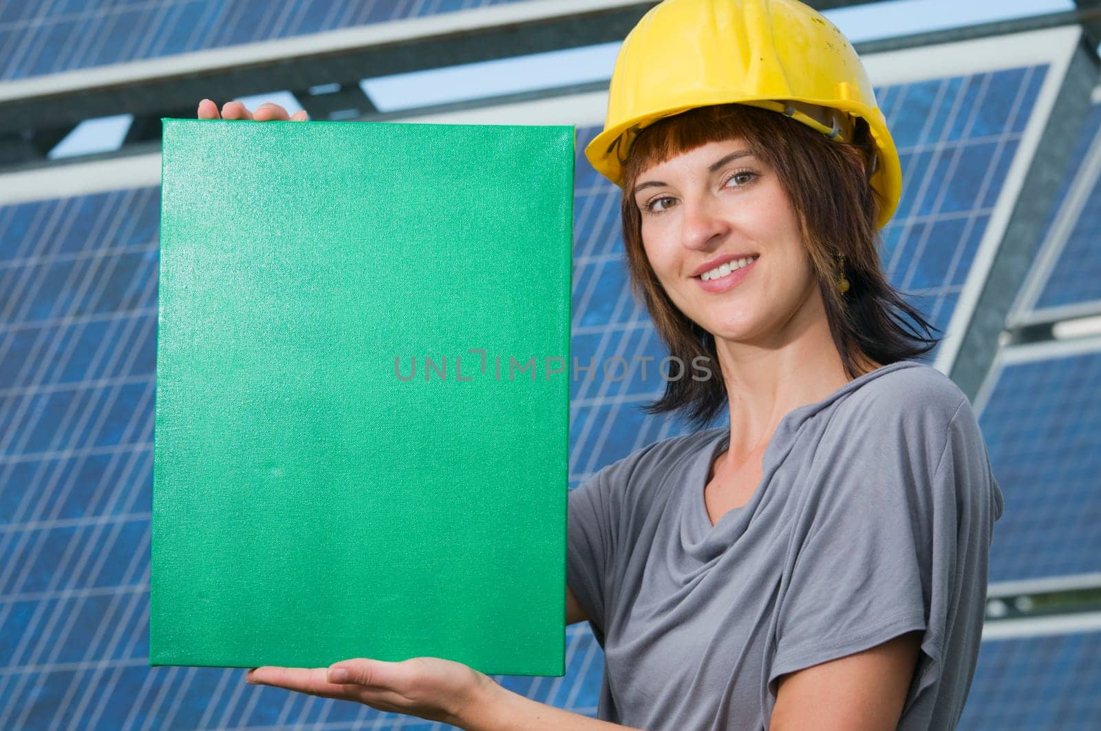 young brunette woman in T-shirt and yellow helmet in front of photovoltaics shows a green area standing sideways smiling into the camera
