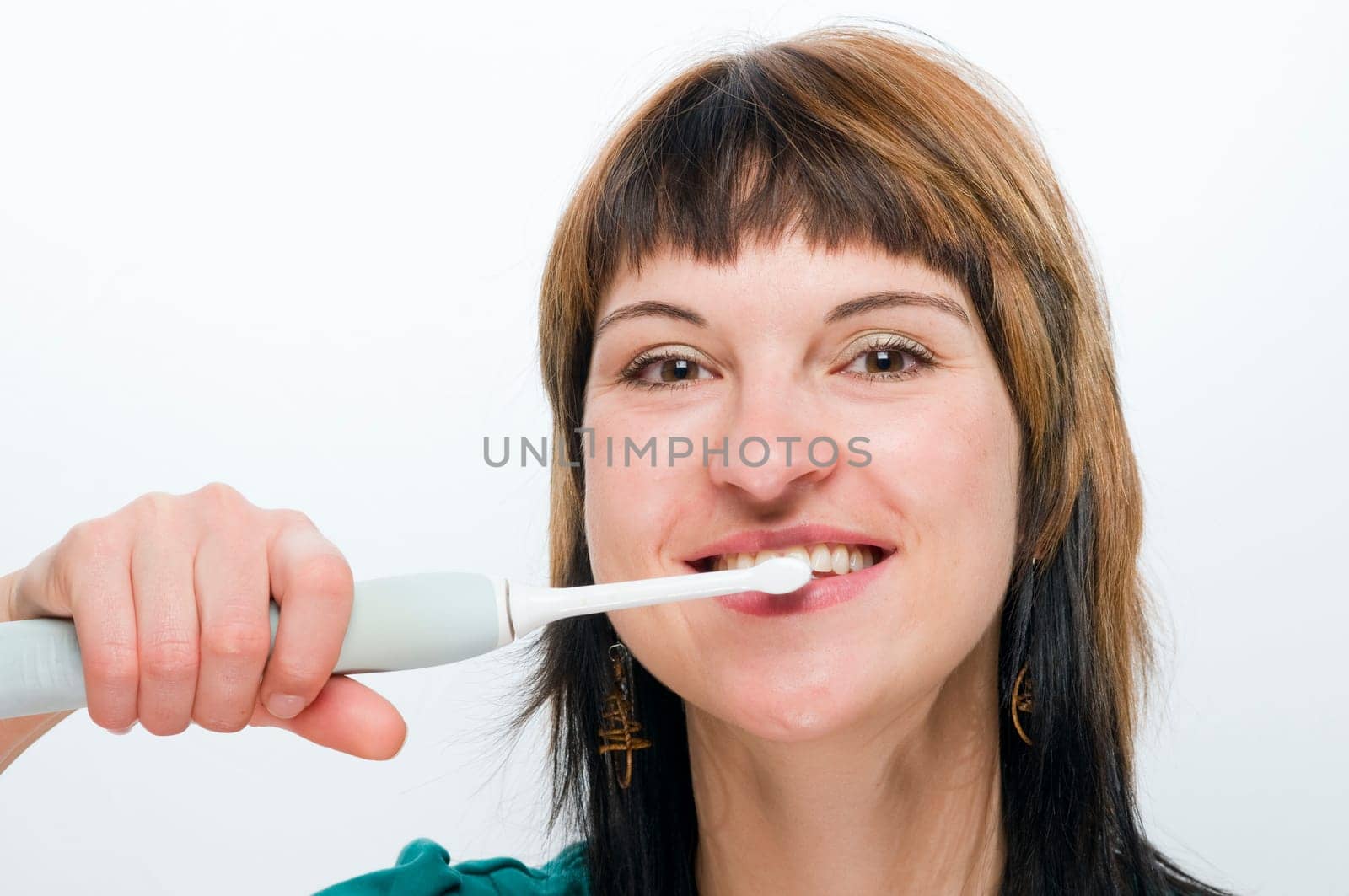 frontal head shot of a young,brunette woman with an electric toothbrush smiling into the camera in front of a white background