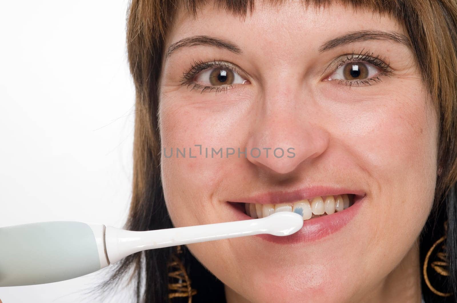 Frontal head shot of young brunette woman with electric toothbrush smiling looking at camera against white background