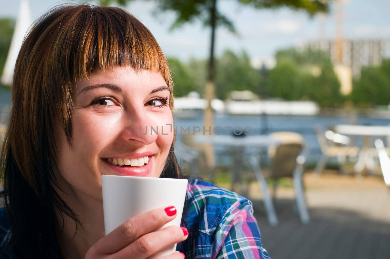 Head-and-shoulder shot of a young,brown-haired woman sitting at a table in the street café,holding a white cup