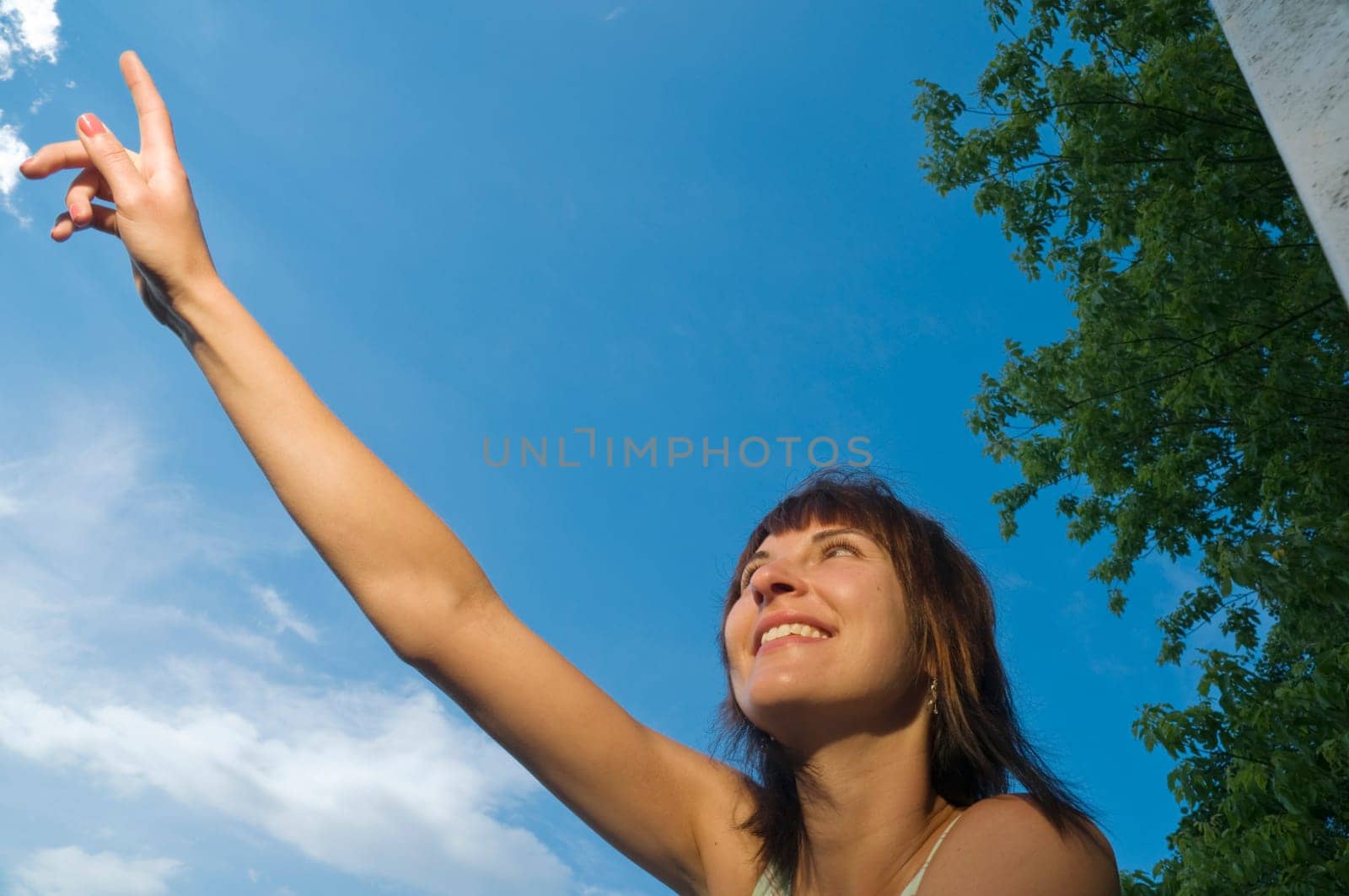Upper body shot of a young,medium-length brown-haired woman in a sleeveless top standing at a lake in Potsdam (Griebnitzsee) and pointing with her right arm nacu upwards in front of a blue sky with small white clouds