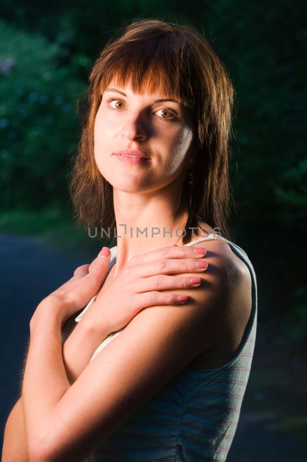 Upper body portrait of a young dark-haired woman in a sleeveless top and crossed arms in front of her chest and hands on her shoulders on a sidewalk in the park at dusk looking into the camera