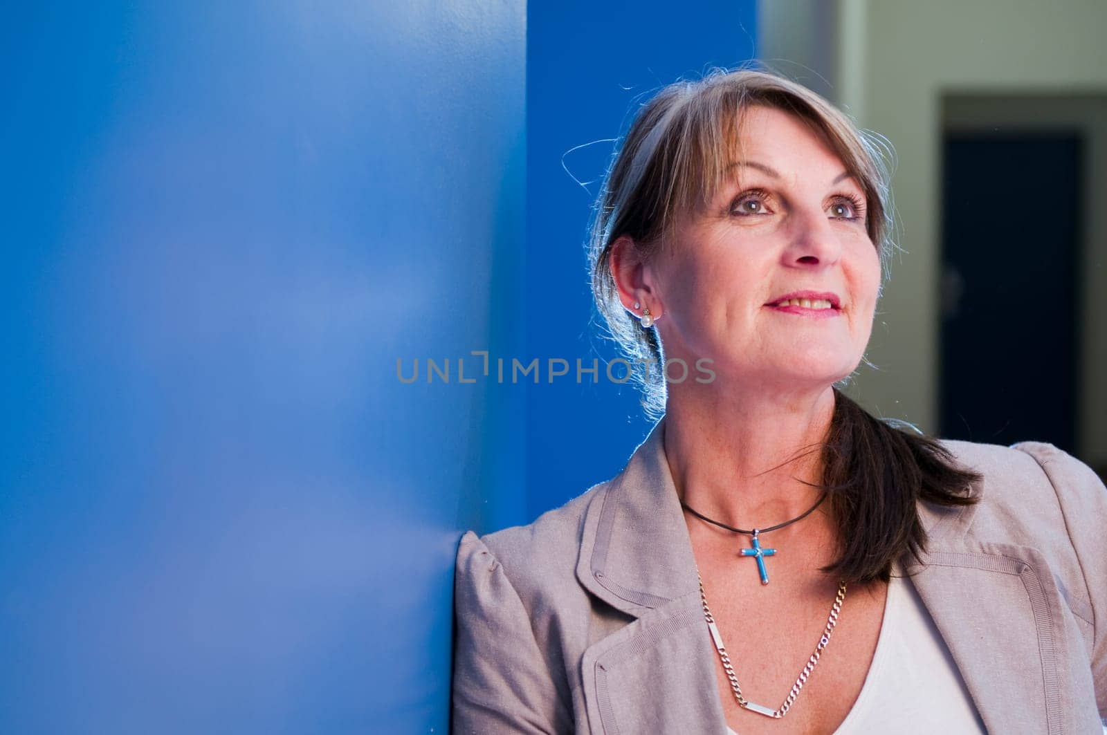 Head-and-shoulder portrait of a middle-aged woman leaning against a blue hallway wall,looking upwards in a friendly manner