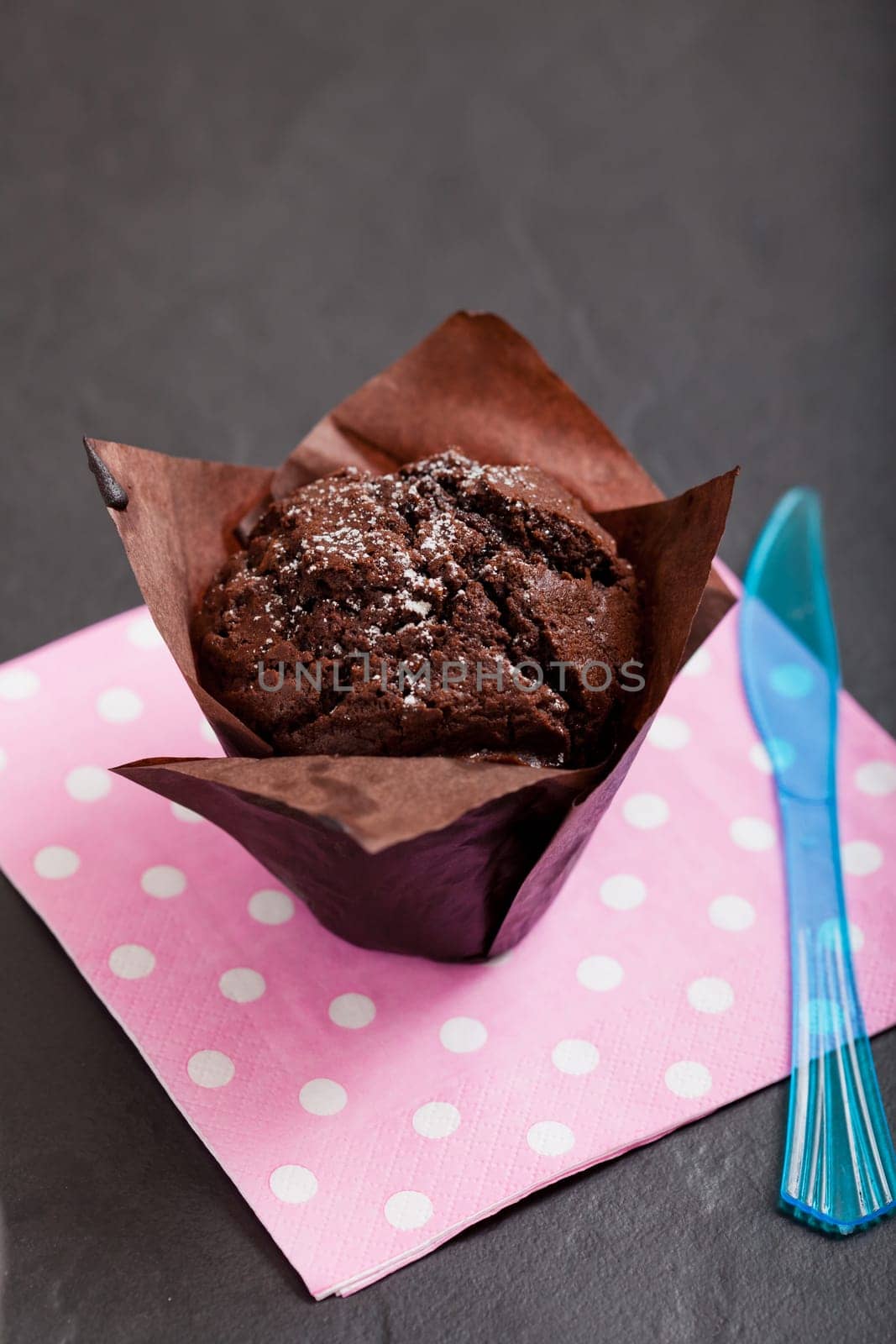 Closeup of chocolate muffin sprinkled with icing sugar and blue plastic fork resting on pink serviette with white spots