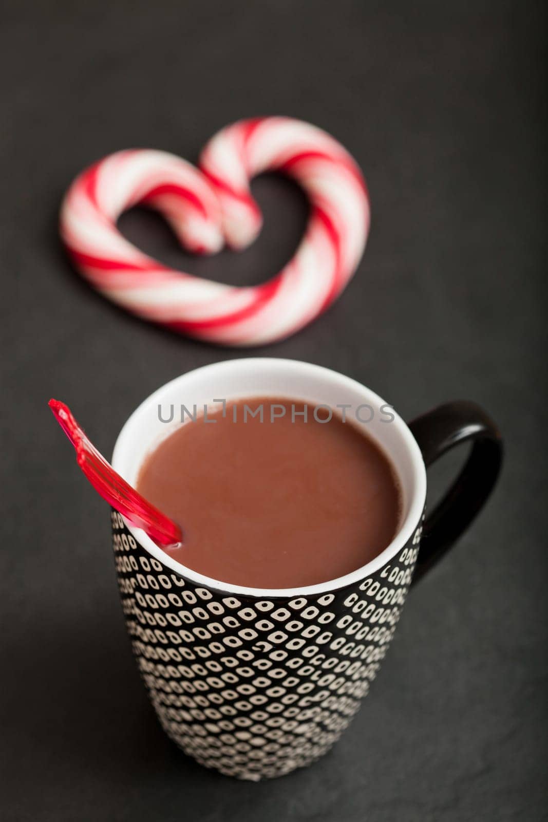 Closeup of mug of hot chocolate with red plastic spoon and hard candy heart with red swirls in background