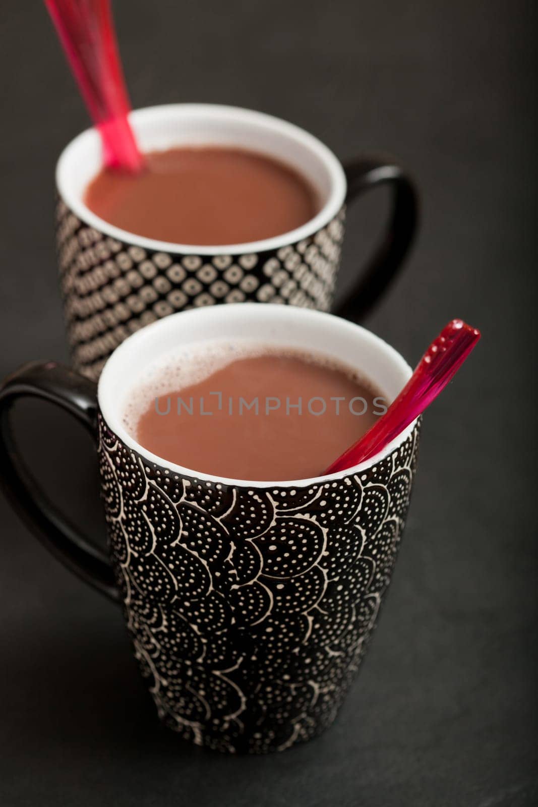 Closeup of two mugs of hot chocolate with red plastic spoons on dark surface