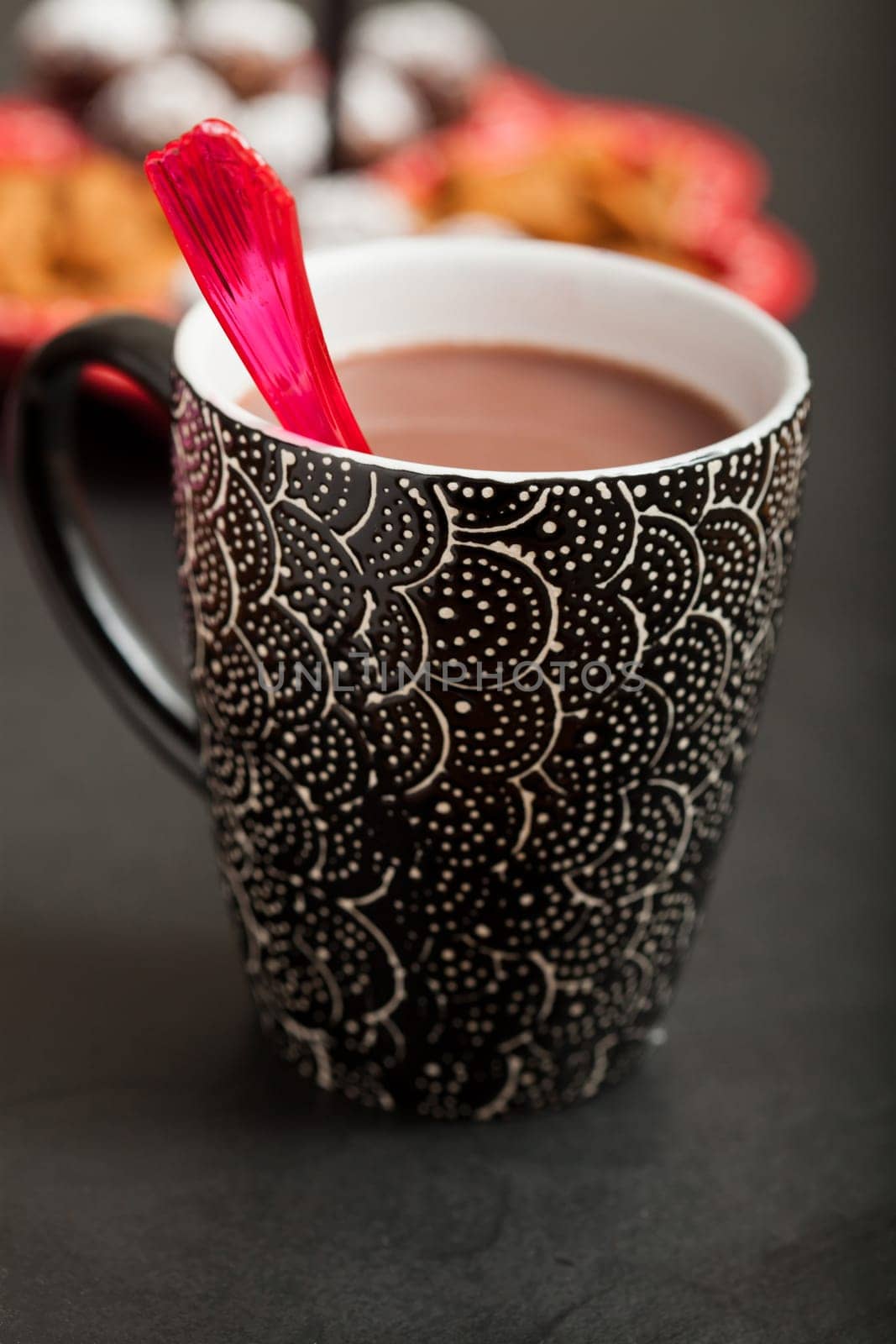 Closeup of mug of hot chocolate with red plastic spoon on dark surface with tray of chocolate balls and almonds in background