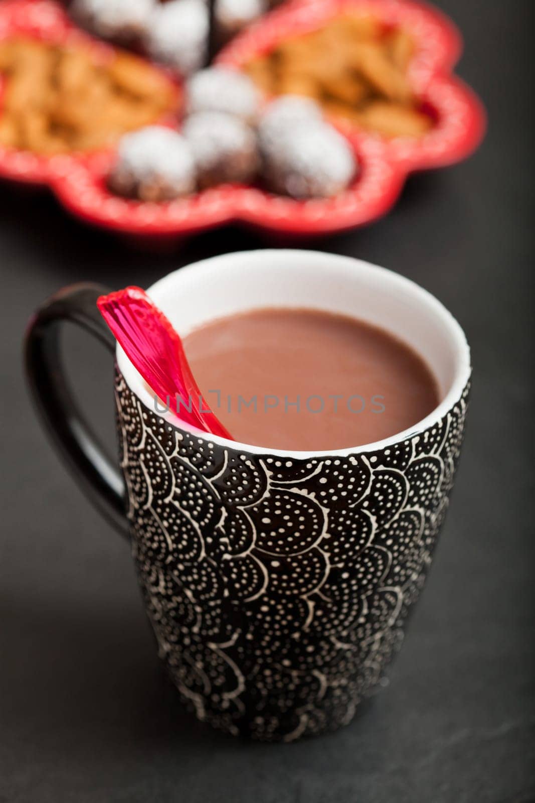 Closeup of mug of hot chocolate with red plastic spoon with tray of chocolate balls and almonds in background