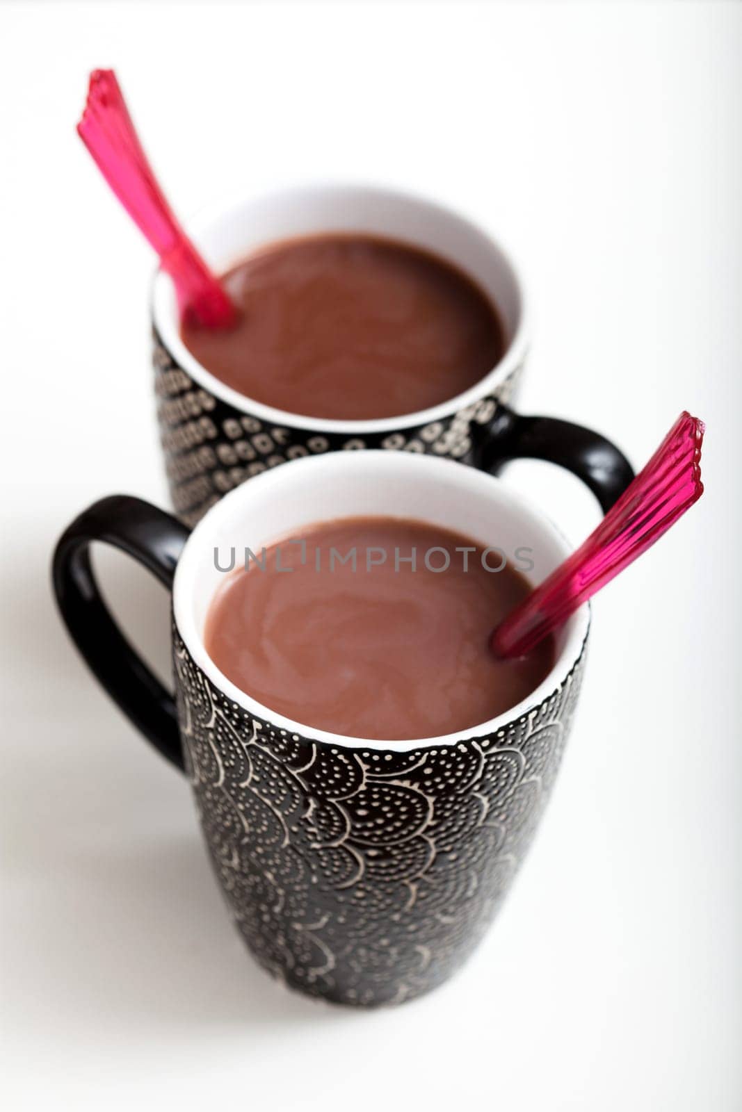 Closeup of two mugs of hot chocolate with red plastic spoons on light surface