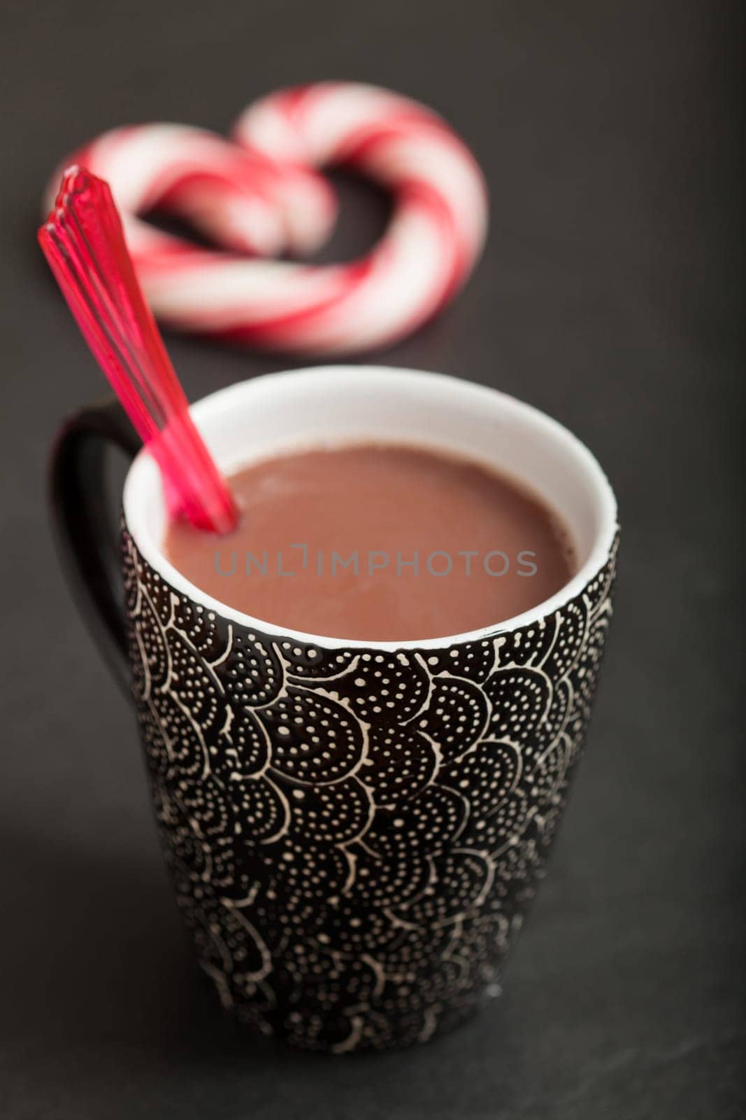 Closeup of mug of hot chocolate with red plastic spoon and hard candy heart with red swirls in background