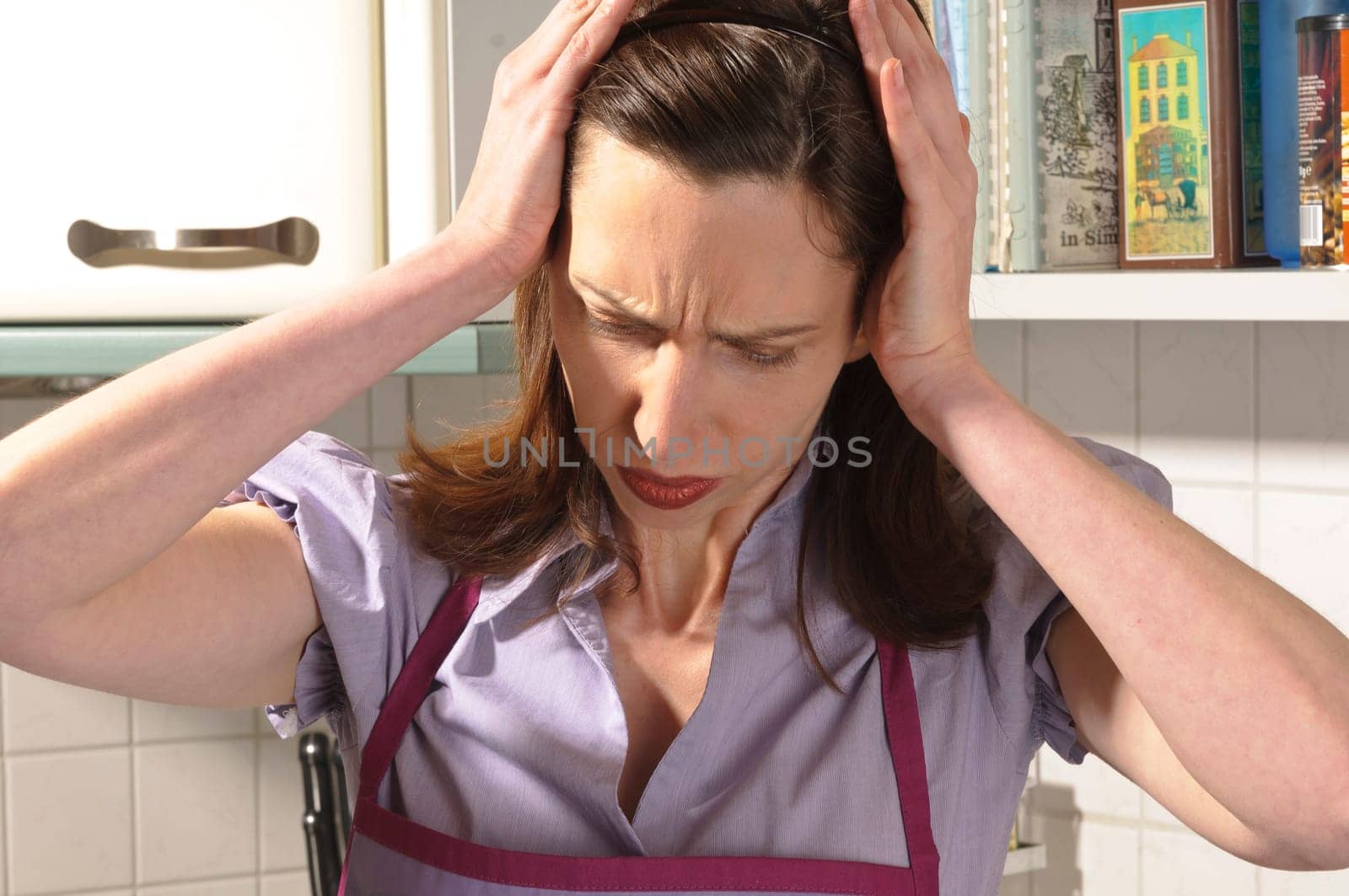 Adult housewife stands in her bright fitted kitchen and holds her hands to her head.