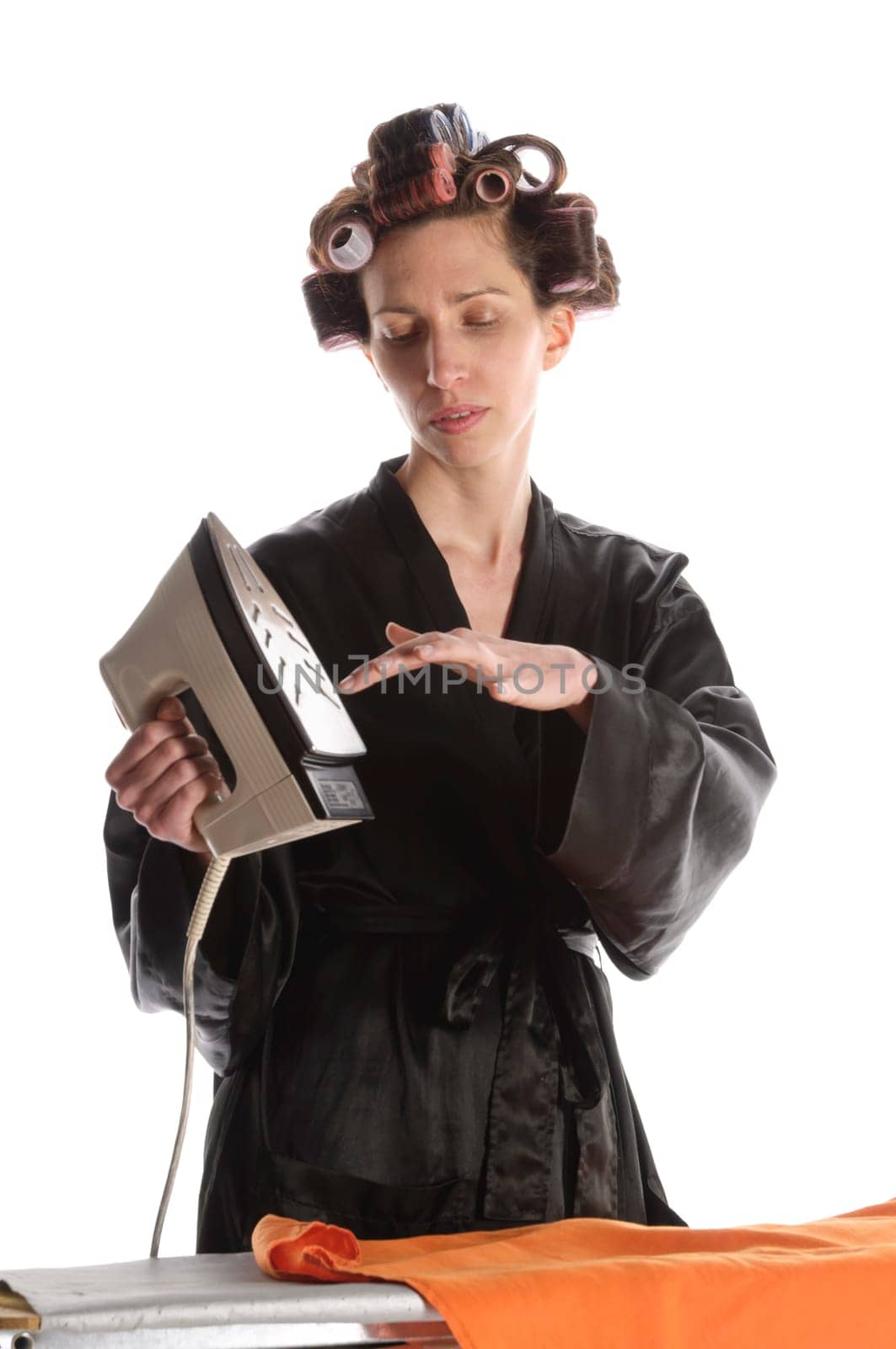 Adult housewife with curlers in her hair and a bathrobe stands behind the ironing board and irons with the iron against a white background.