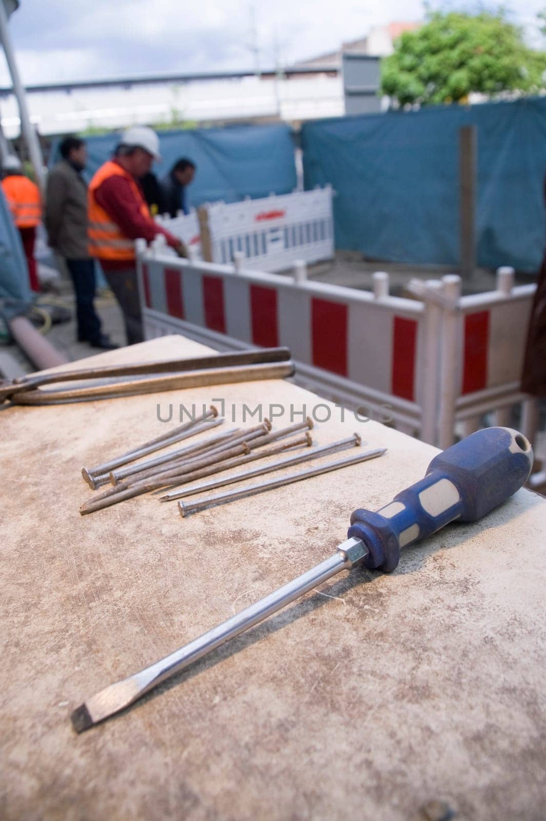 Wide-angle close-up of a screwdriver on a table with nails on a construction site with blurred and unrecognizable construction workers in the background in daylight
