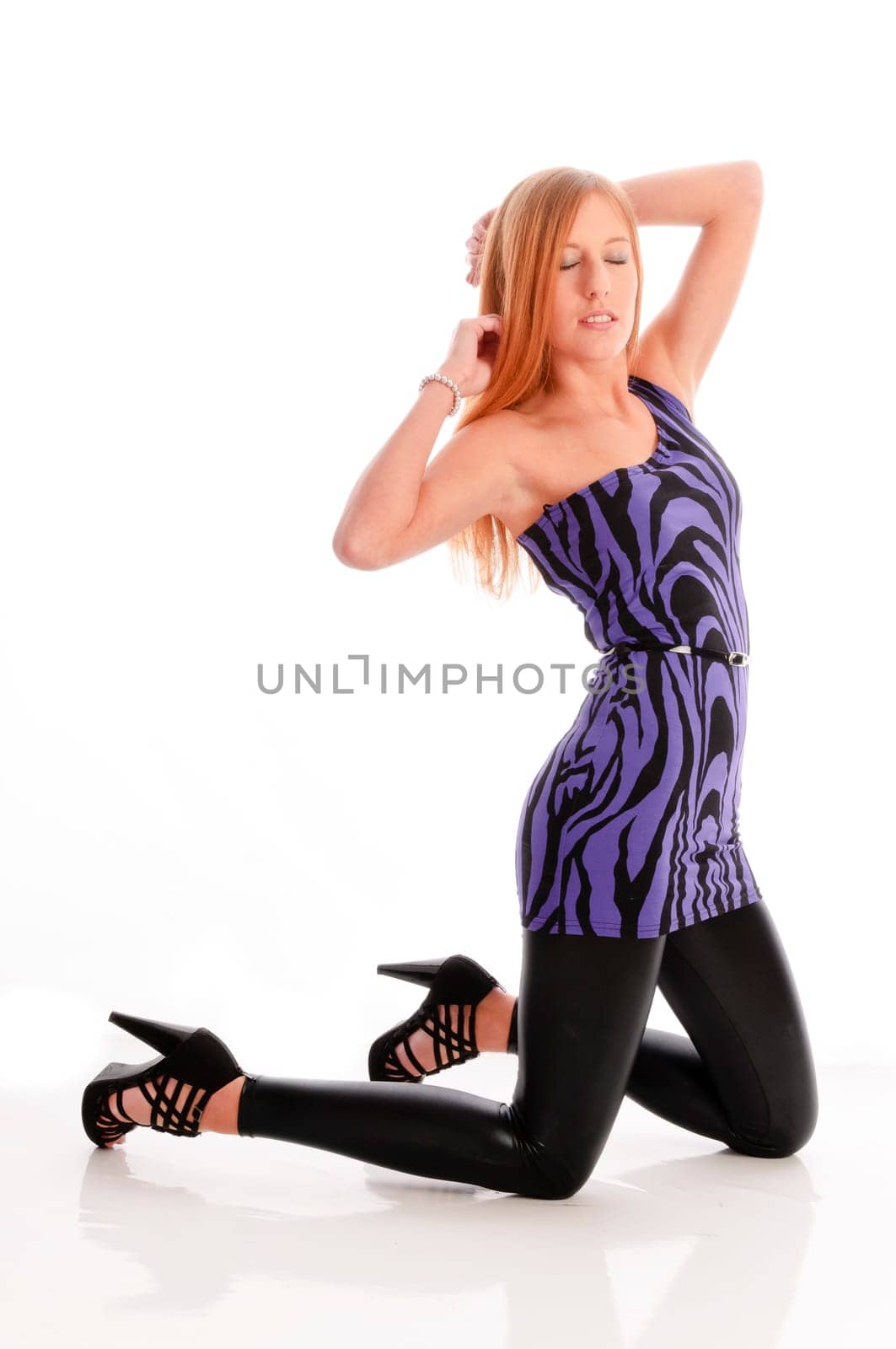 Attractive young woman with long blonde hair wears a blue mini dress with black glossy leggings and poses isolated against a white background.