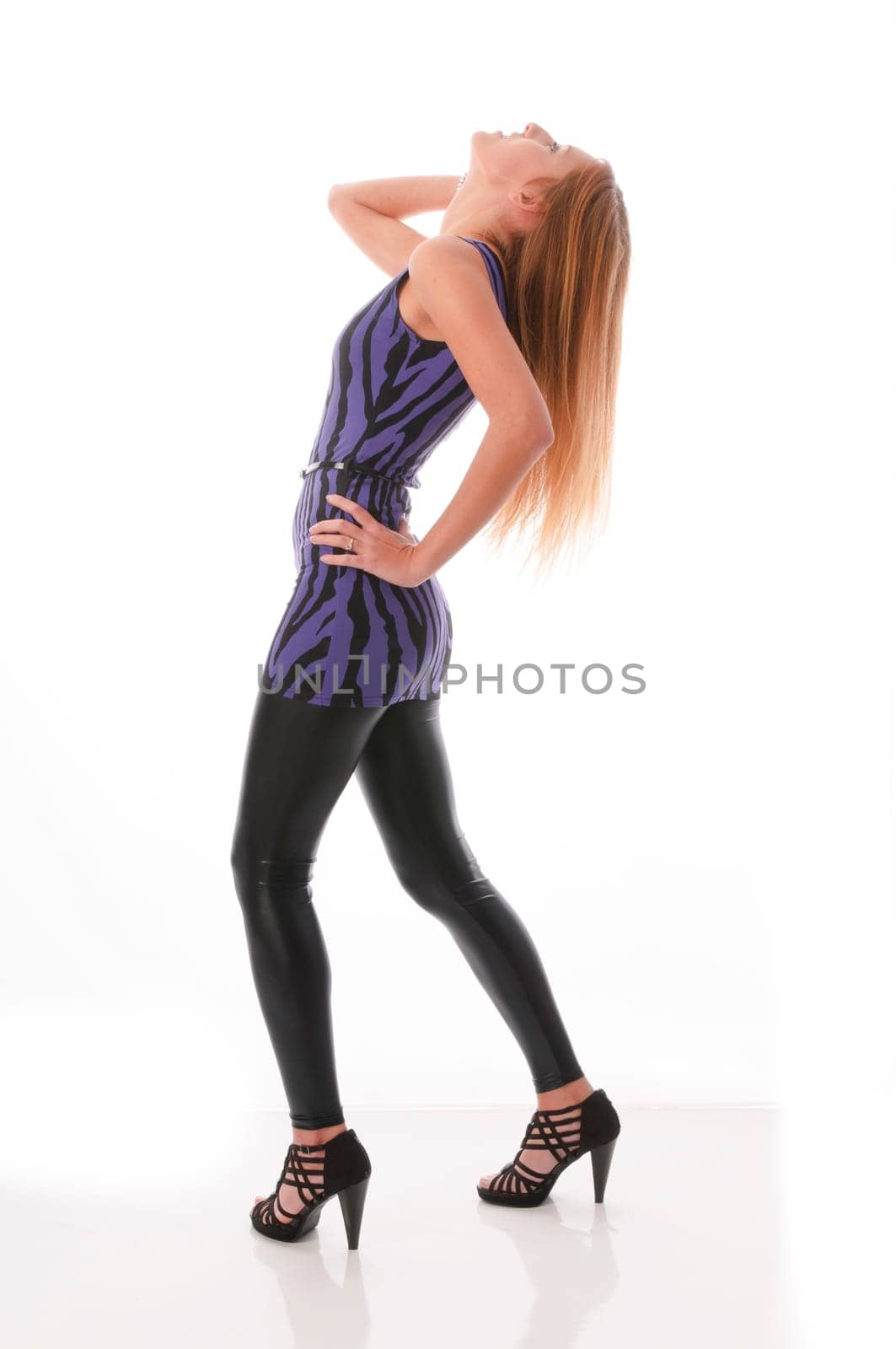 Attractive young woman with long blonde hair wears a blue mini dress with black glossy leggings and poses isolated against a white background.
