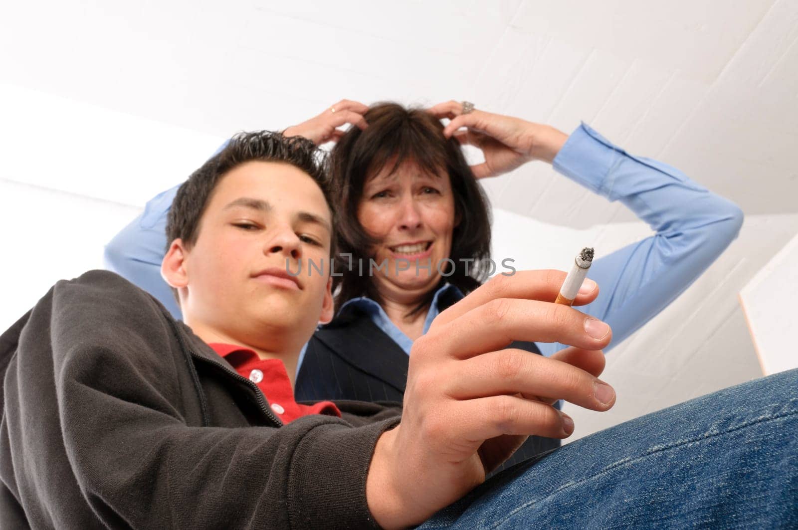 The boy sits casually in the room and smokes,the mother stands horrified behind it,in front of a white background.