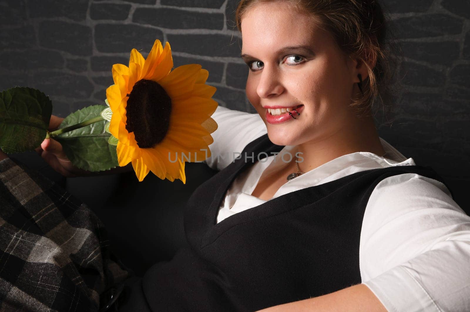 A young girl with a blouse and boots sits teasingly on a black leather armchair against a dark background and plays with a sunflower