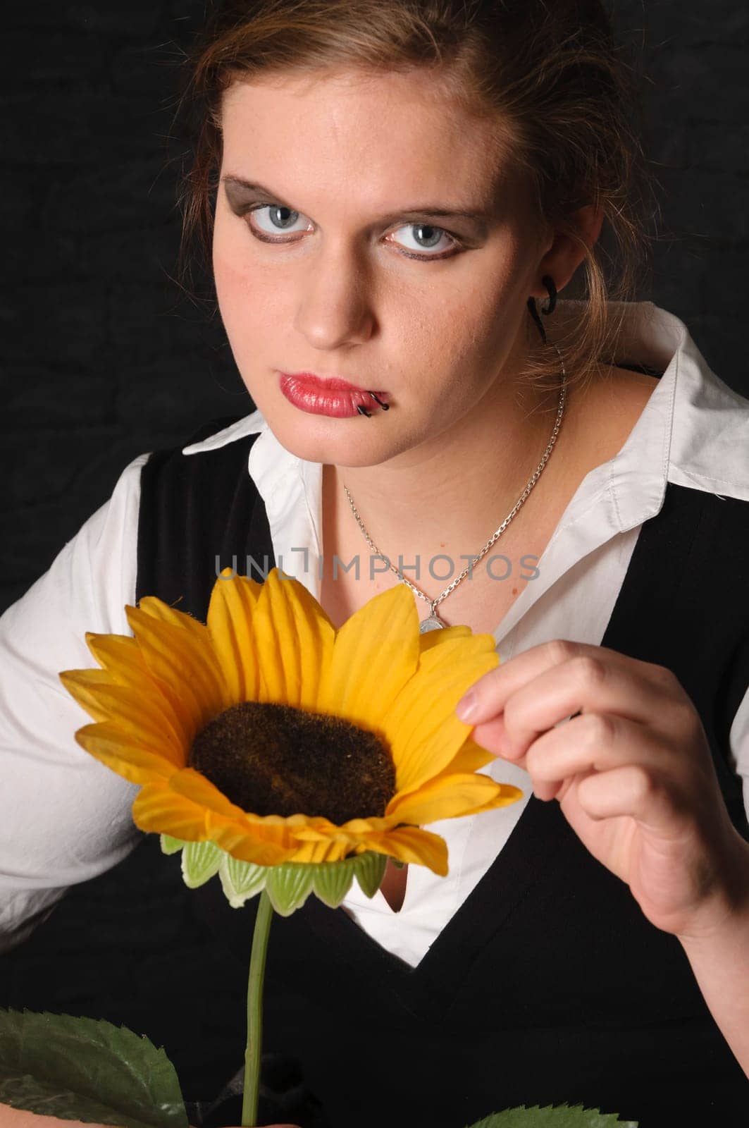 A young girl with a blouse and boots sits teasingly on a black leather armchair against a dark background and plays with a sunflower