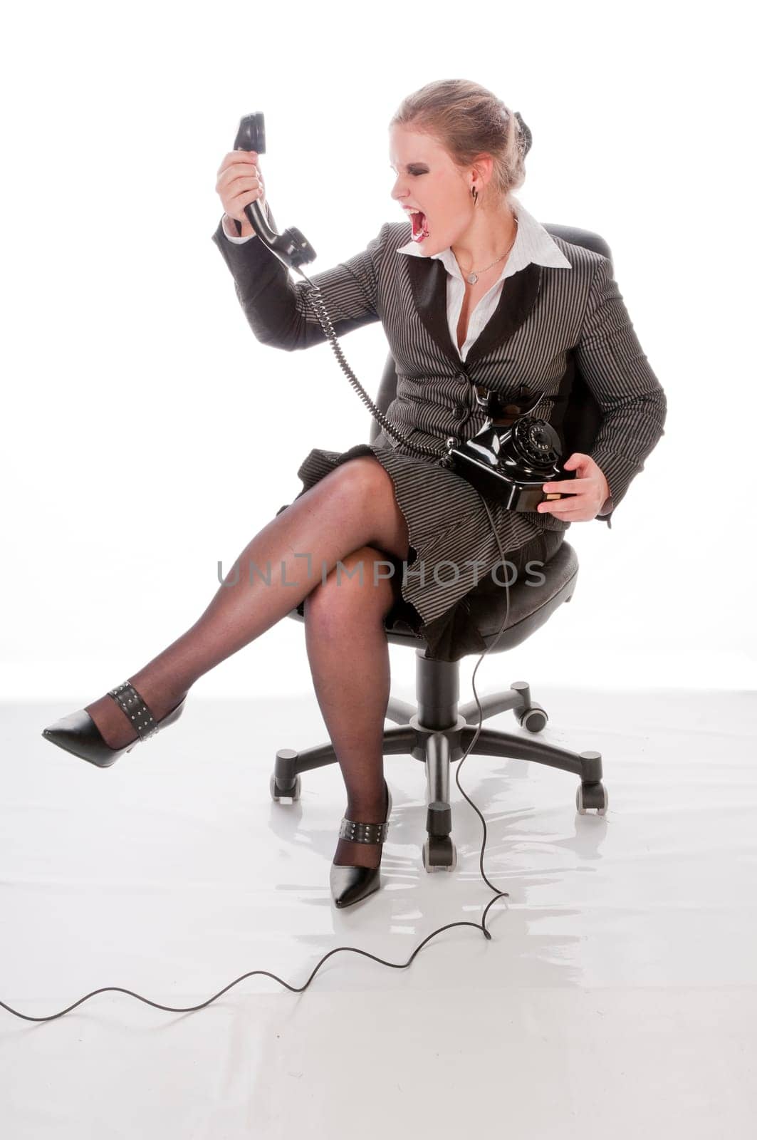 Young woman in grey business suit and high heels makes calls with landline telephone and yells angrily into the receiver against a white background